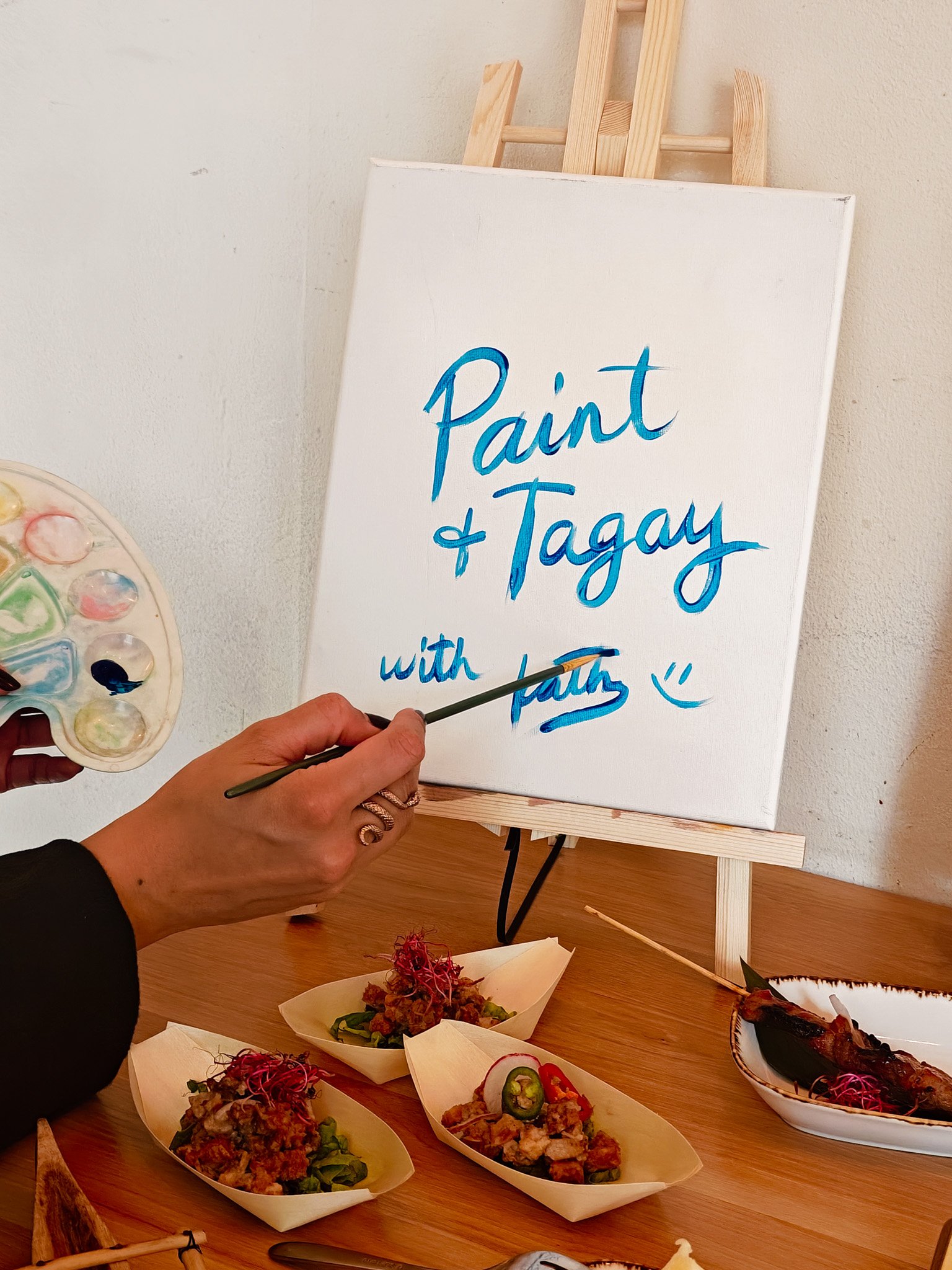 A person is painting the words "Paint & Tagay with faith :)" on a canvas placed on an easel. The table is set with a palette, paint, and various small dishes of food, including skewers and other garnished items.