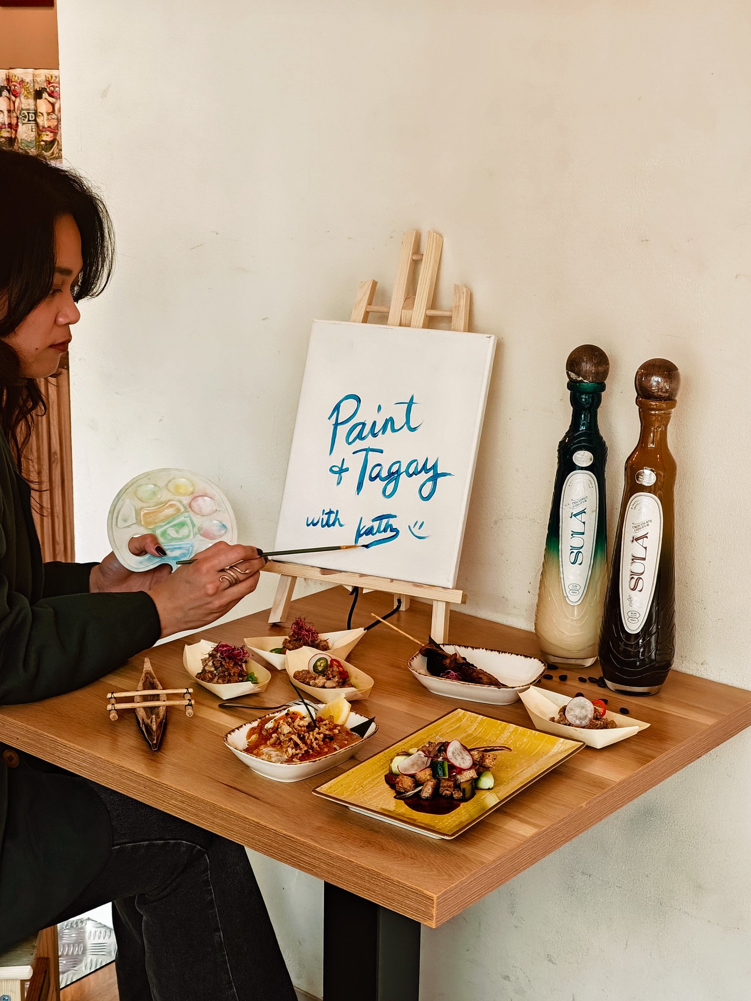 Person painting at a table with an art easel, paint palette, small bowls of food, and two bottles on display, labeled with "Paint & Tagay with Kath."
