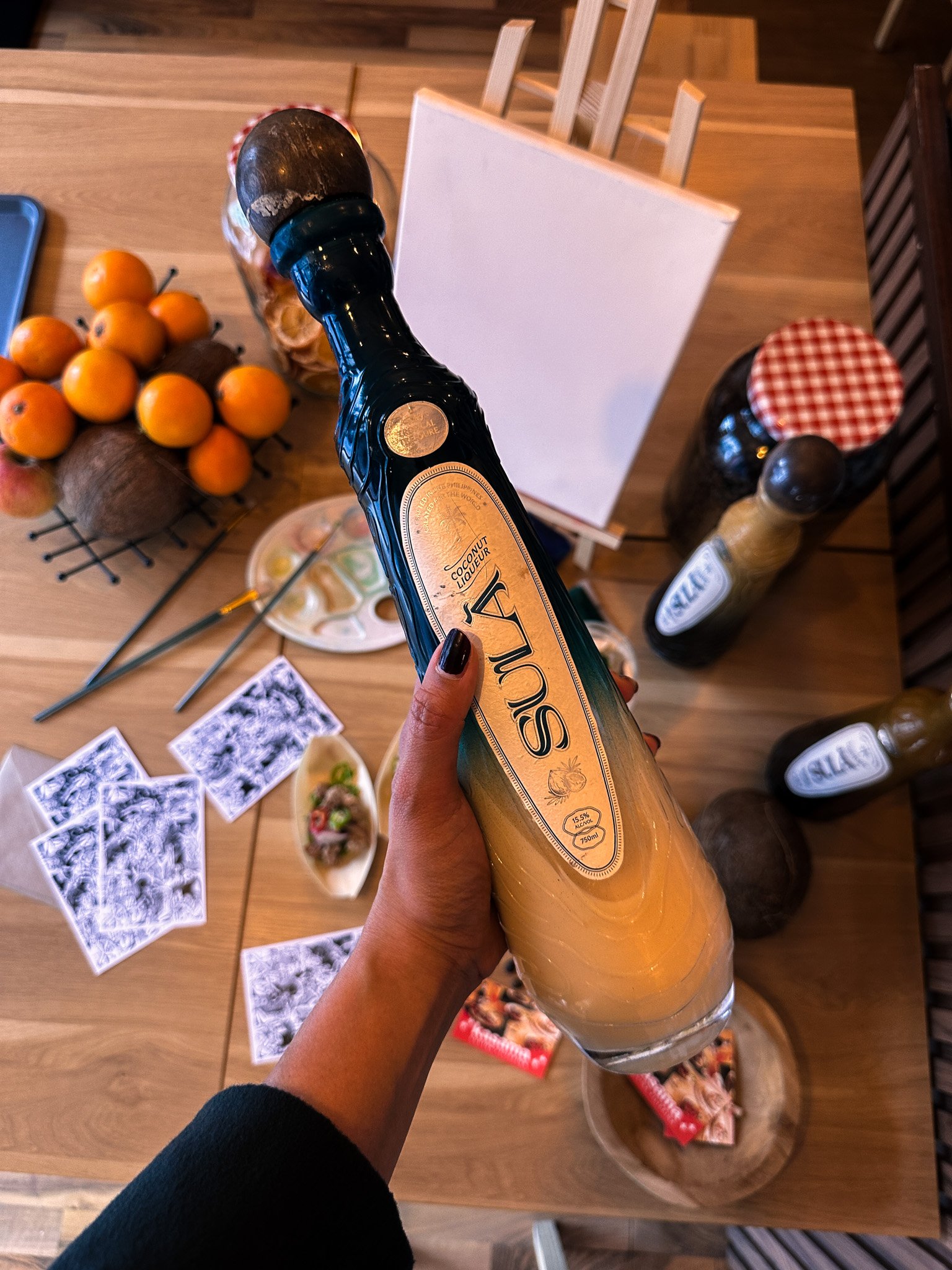 Hand holding Slua coconut liqueur bottle over a table with paint supplies, coconuts, oranges, and printed cards.