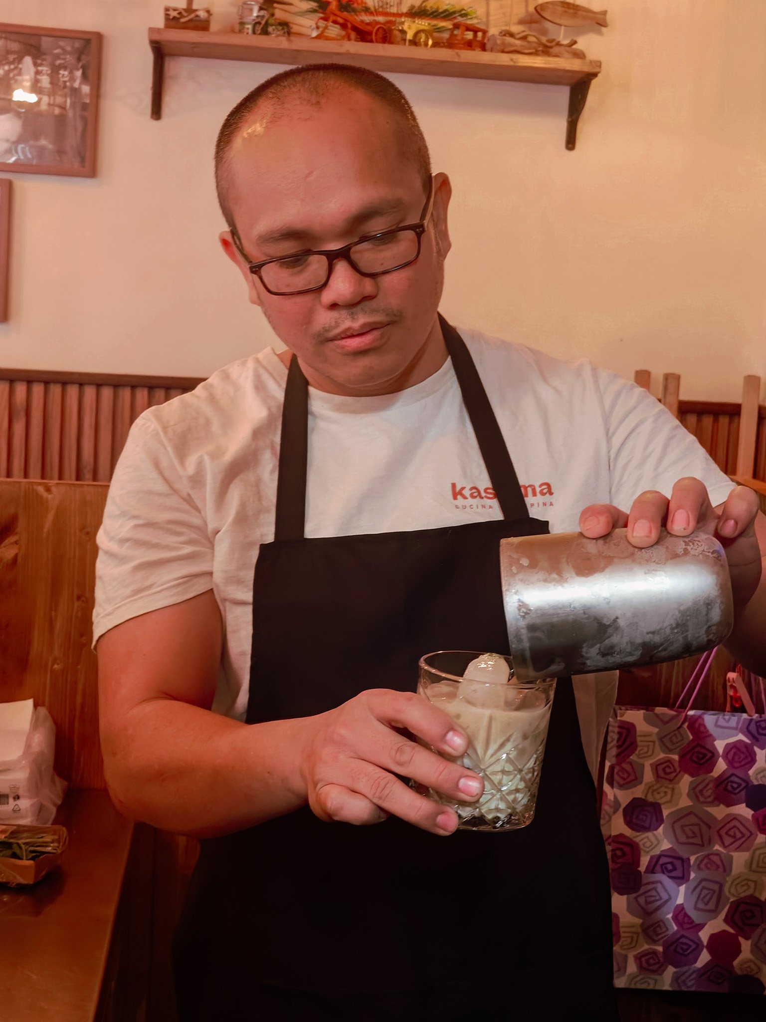 Person in a white shirt and black apron pouring a beverage into a glass with ice inside a cozy, decorated room.