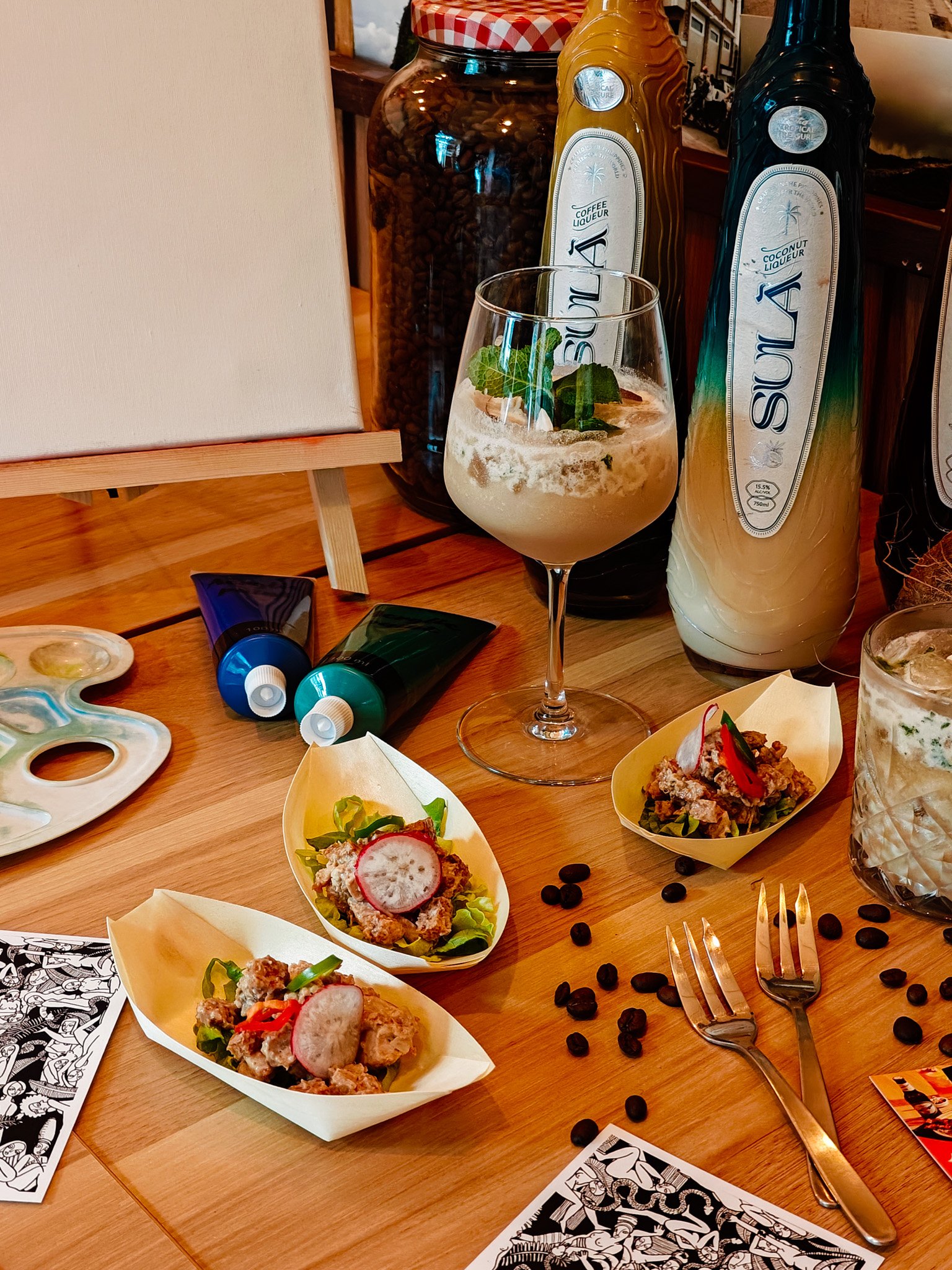Artistic table setting with coffee liqueur bottles, a cocktail glass, food on wooden plates, paint tubes, and scattered coffee beans.