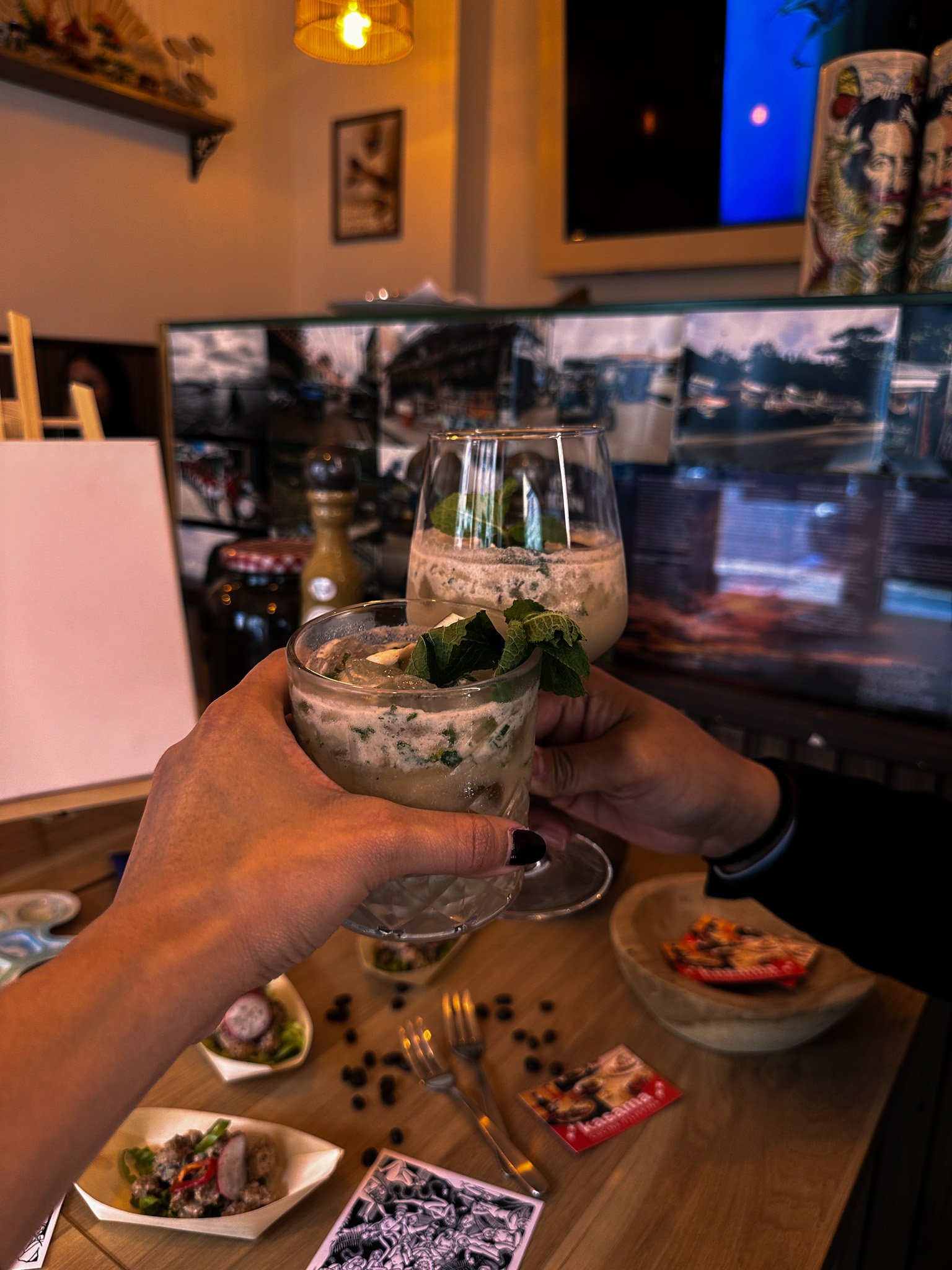 Two people toasting with creamy cocktails garnished with mint, surrounded by small dishes of food, coffee beans, and cards on a wooden table in a cozy setting.