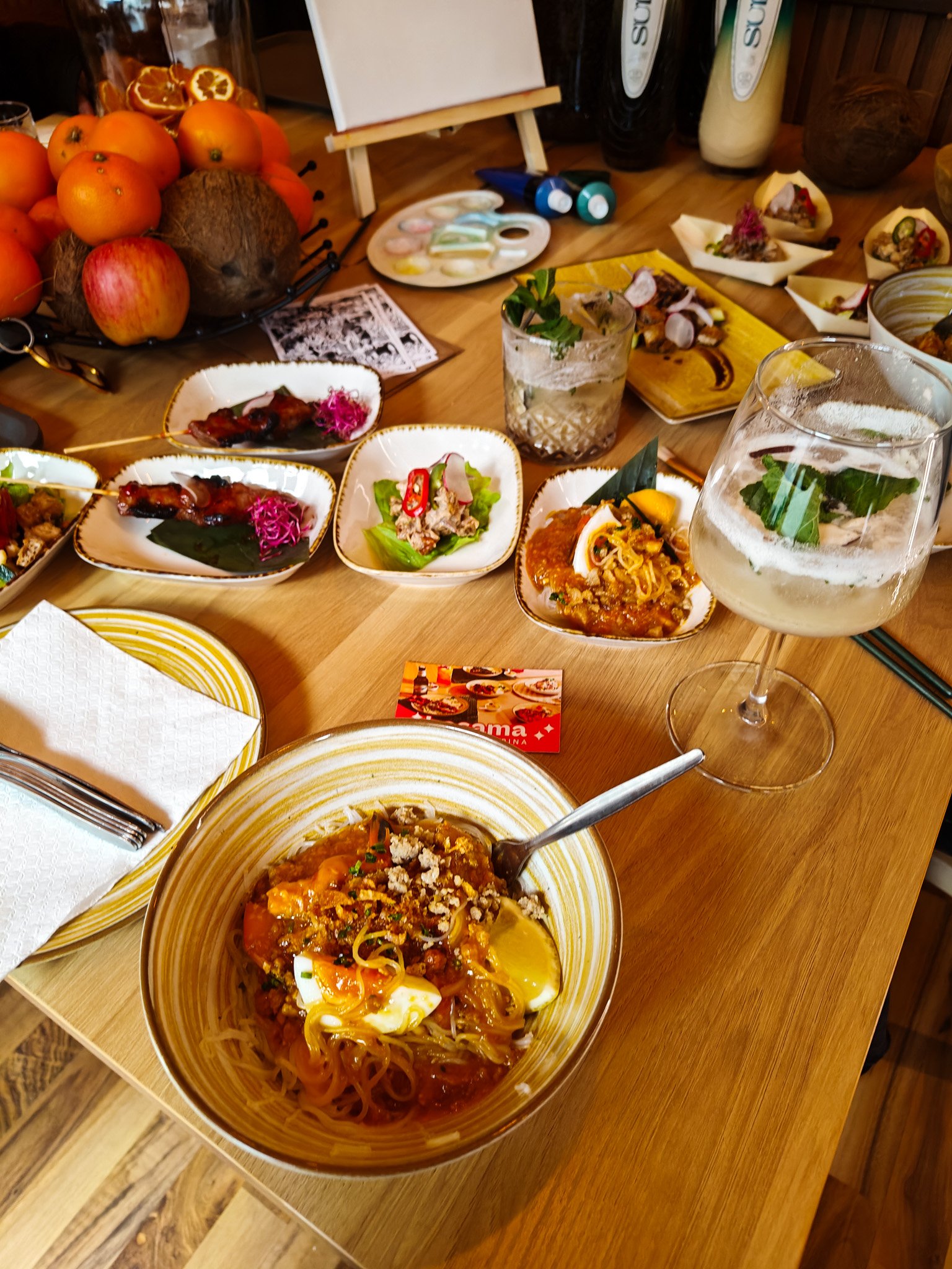 A table set with various dishes including a bowl of pasta with sauce and lemon, small plates with appetizers, and a glass of white wine. There are also decorative items like oranges and coconuts, paint tubes, and a canvas in the background.