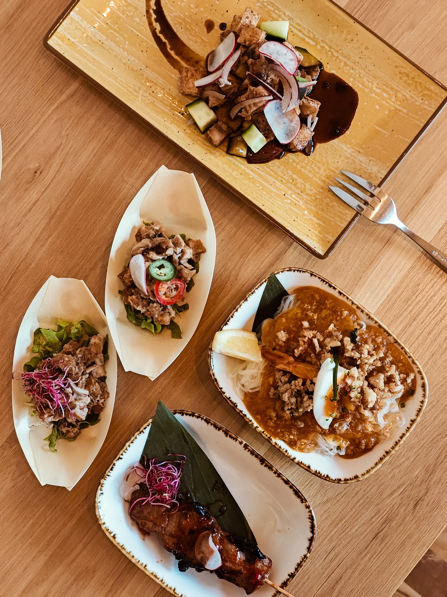 Assorted Asian dishes on wooden table, including skewers, salad with radish, noodle dish with egg and ground meat, served in decorative plates.