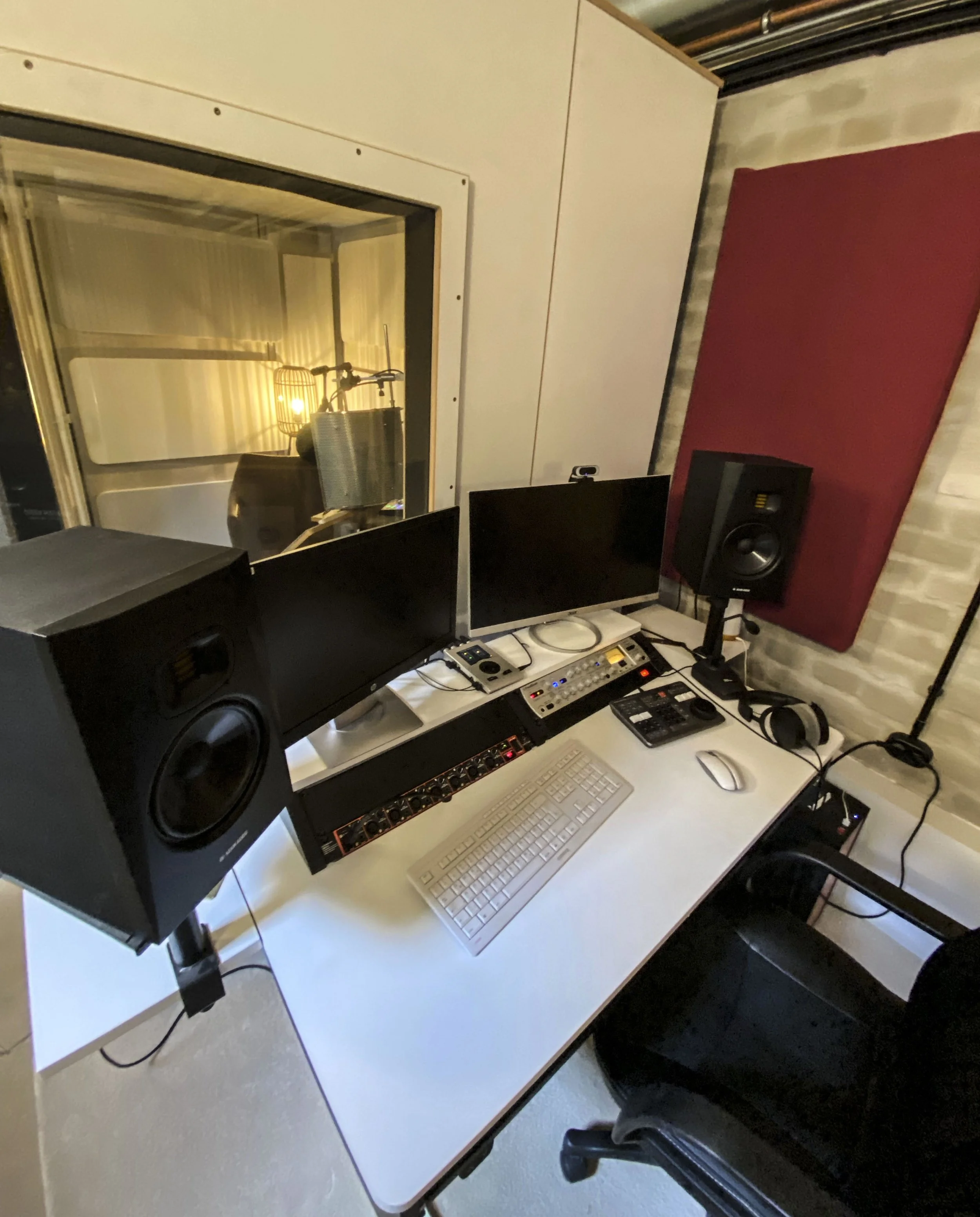 Aufnahme eines Audio- oder Musikproduktionsraums mit Monitorlautsprechern, einem Computer, einem MIDI-Controller, einem Mischpult, einem Keyboard, einem Monitor, einer roten Akustikmatte und einem Mikrofon im Schaltraum in der Hintergrund. 