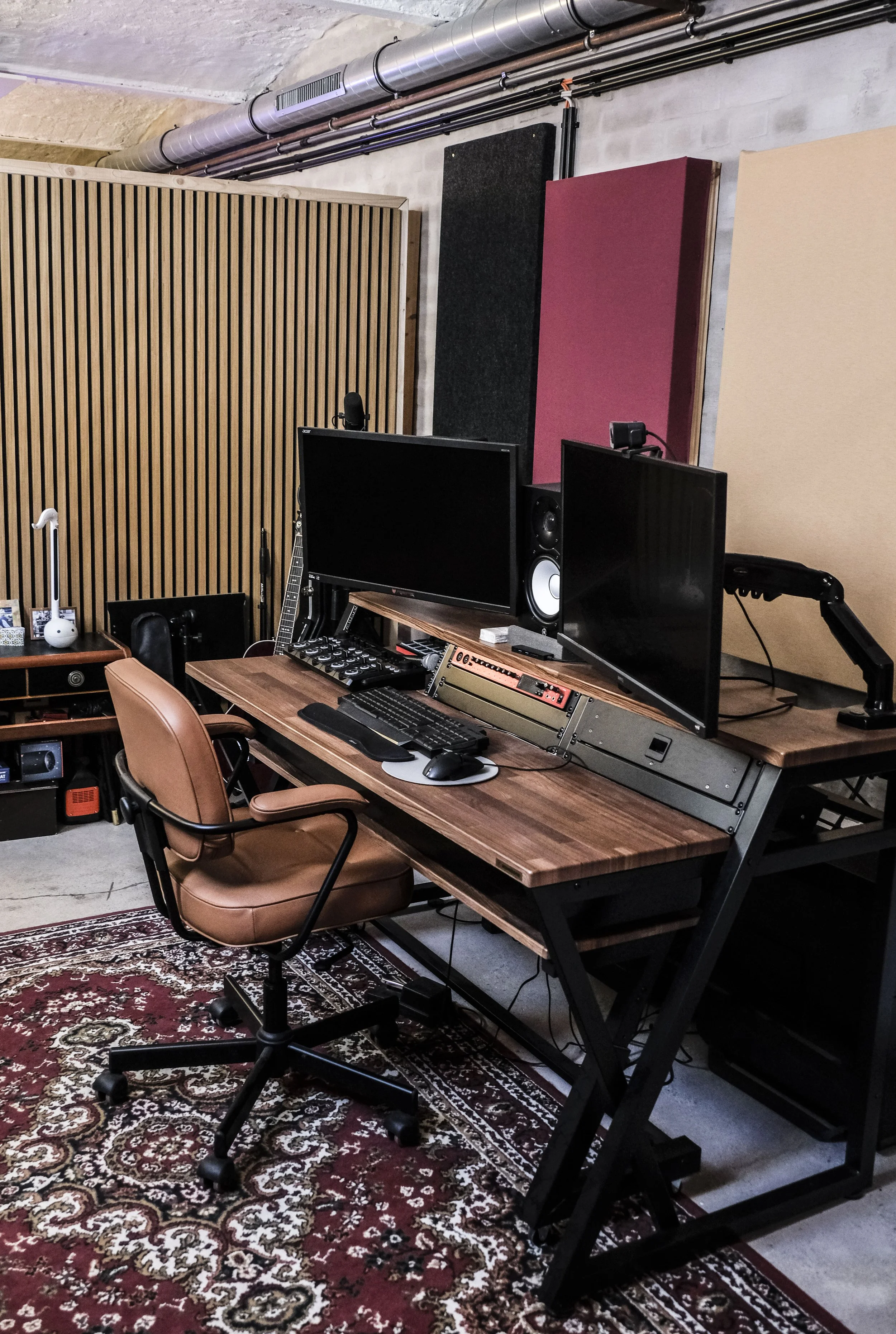 Aufnahme eines Musikproduktionsstudios mit einem großen Holztisch, mehreren Monitoren, einem elektronischen Mischpult, einem Stuhl, Akustikschaumstoffplatten an der Wand, Holzpaneelen und technischen Geräten, in einem Raum mit Betonwänden und sichtbaren Rohrleitungen.