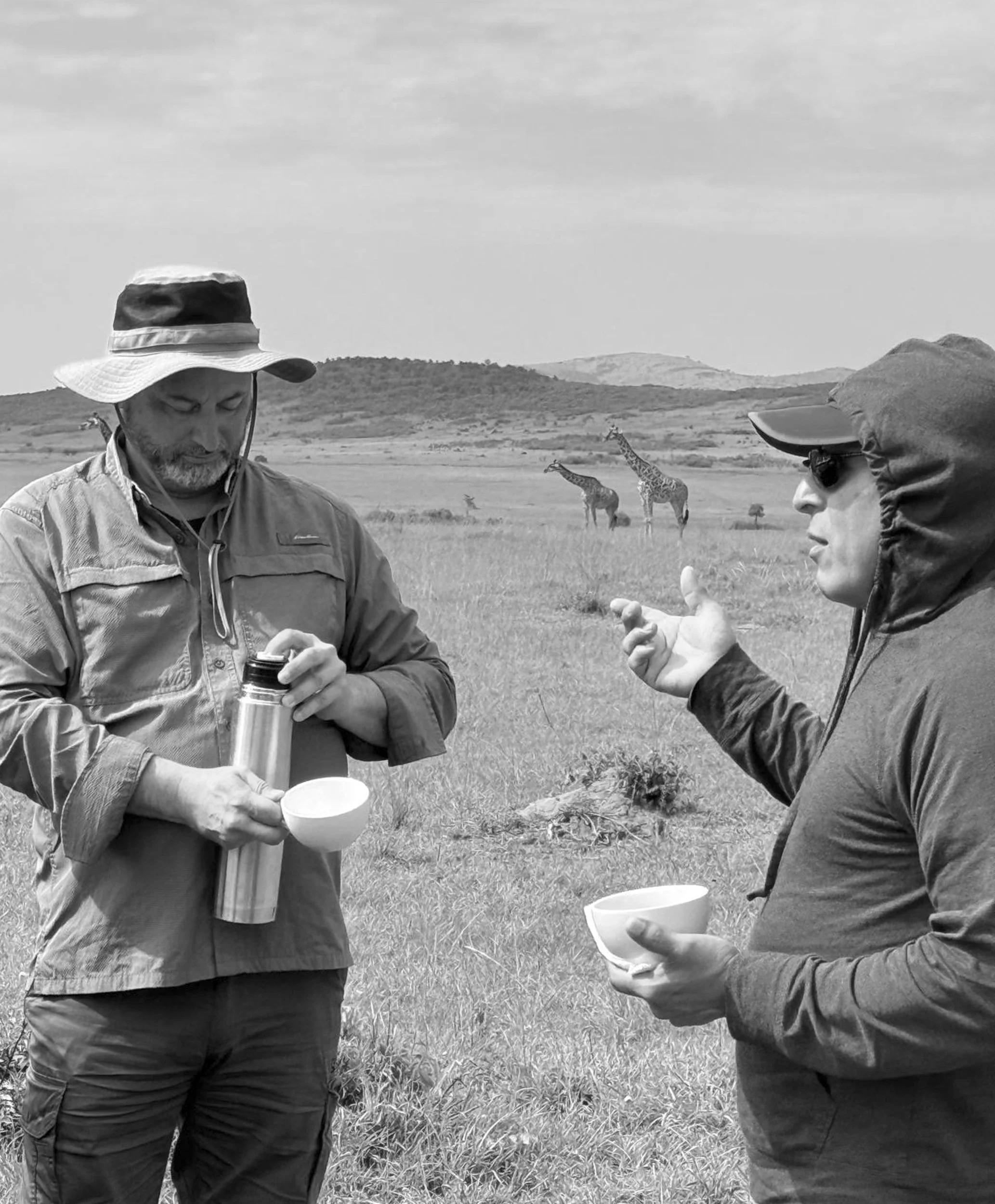 Two men standing in a savannah with giraffes in the background, holding cups and engaging in conversation.