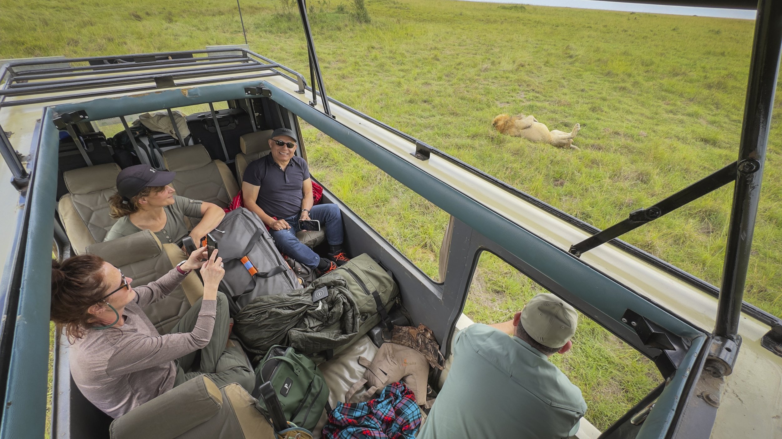 Tourists sitting inside an open-air safari vehicle, observing a male lion lying on the grass outside.