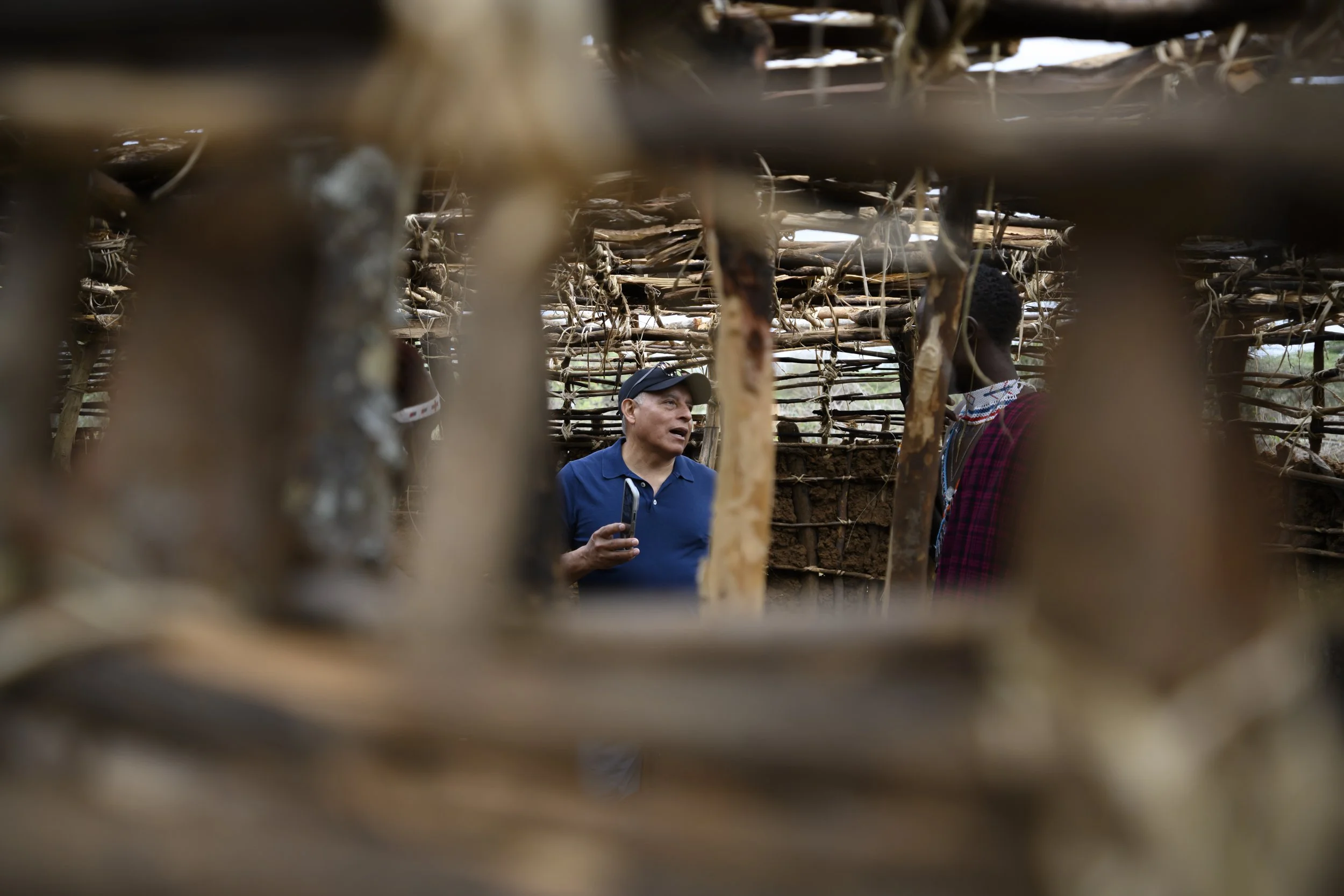 A person wearing a baseball cap speaking to a person in traditional clothing inside a rustic hut made of sticks.