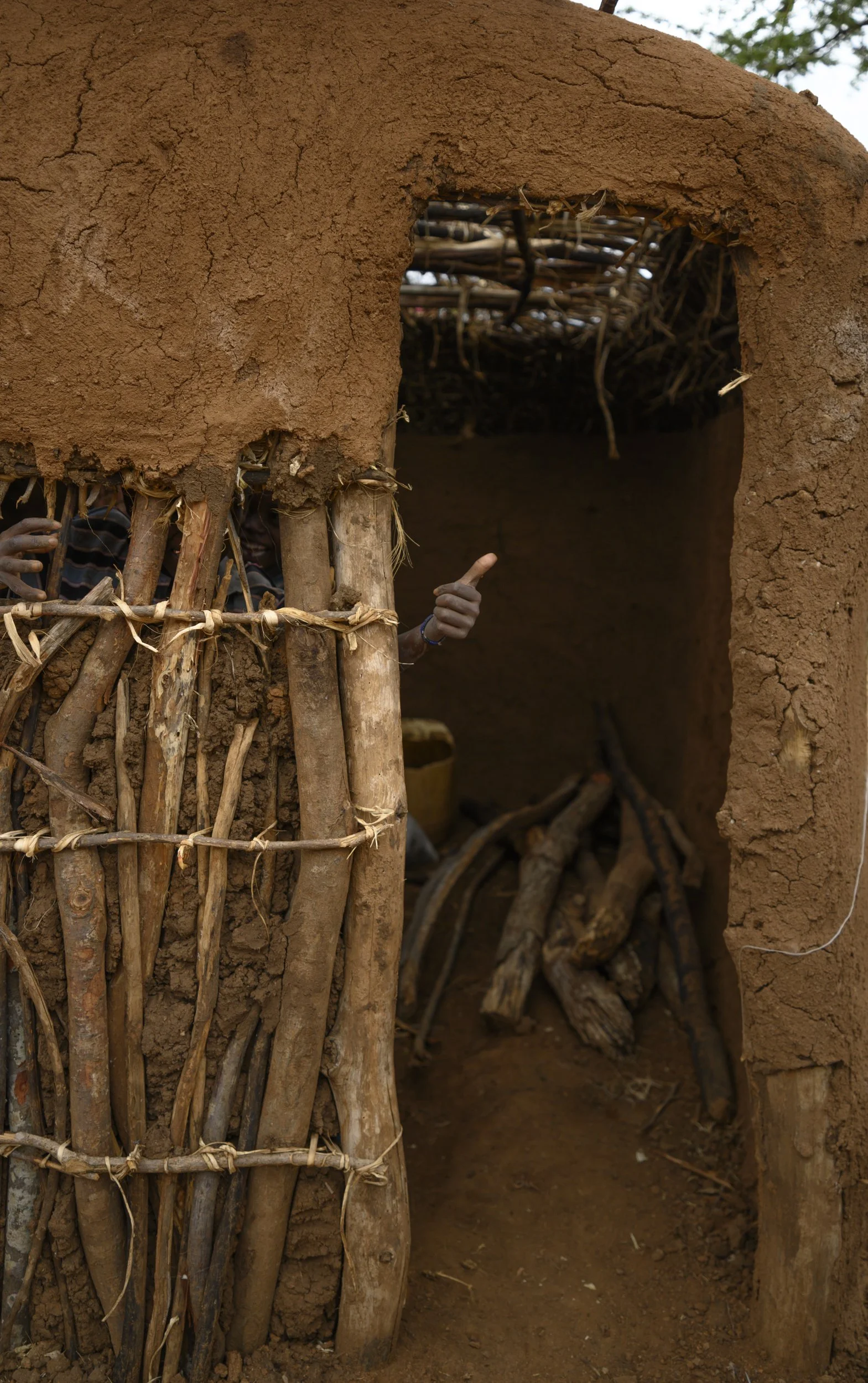 Close-up of a traditional mud-and-stick hut with a small opening, showing a person giving a thumbs-up inside.