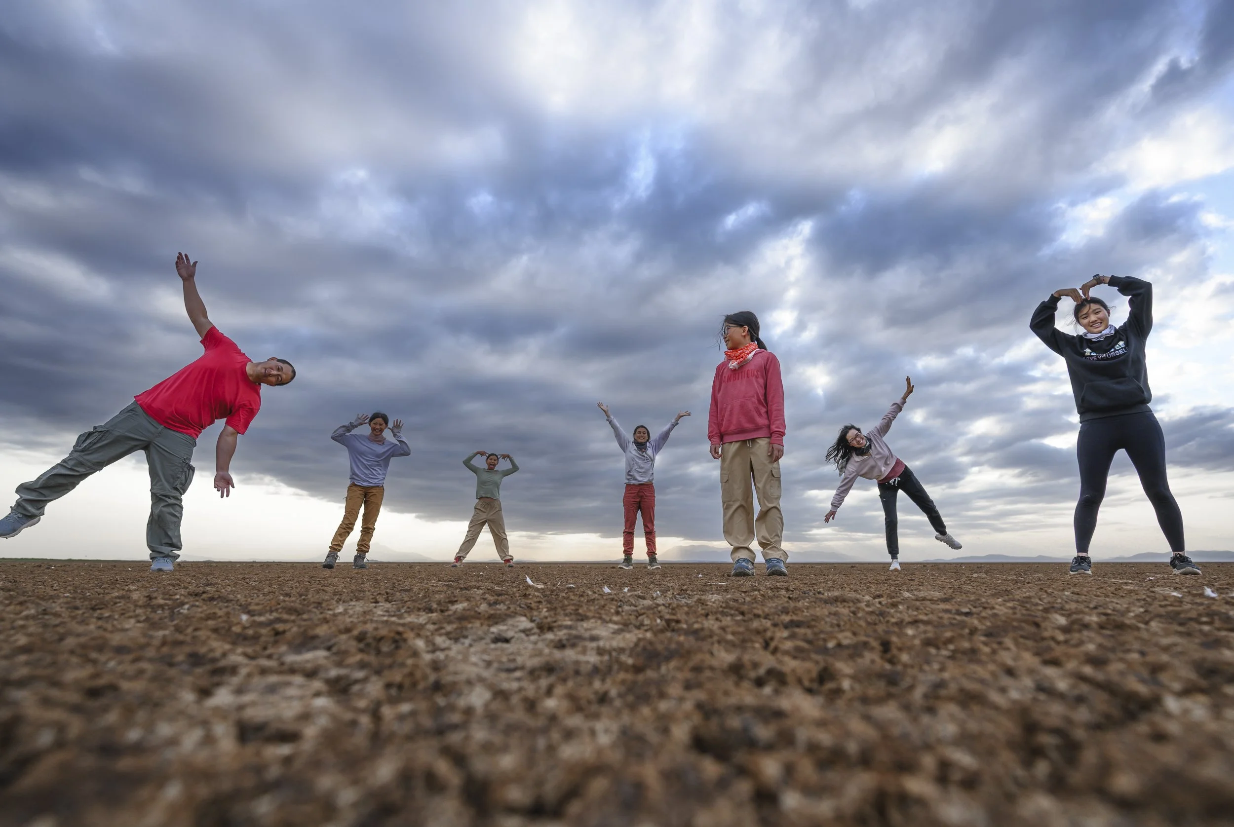 Group of people standing on a dirt field outdoors, some with arms raised, with a cloudy sky overhead, engaging in a joyful activity.
