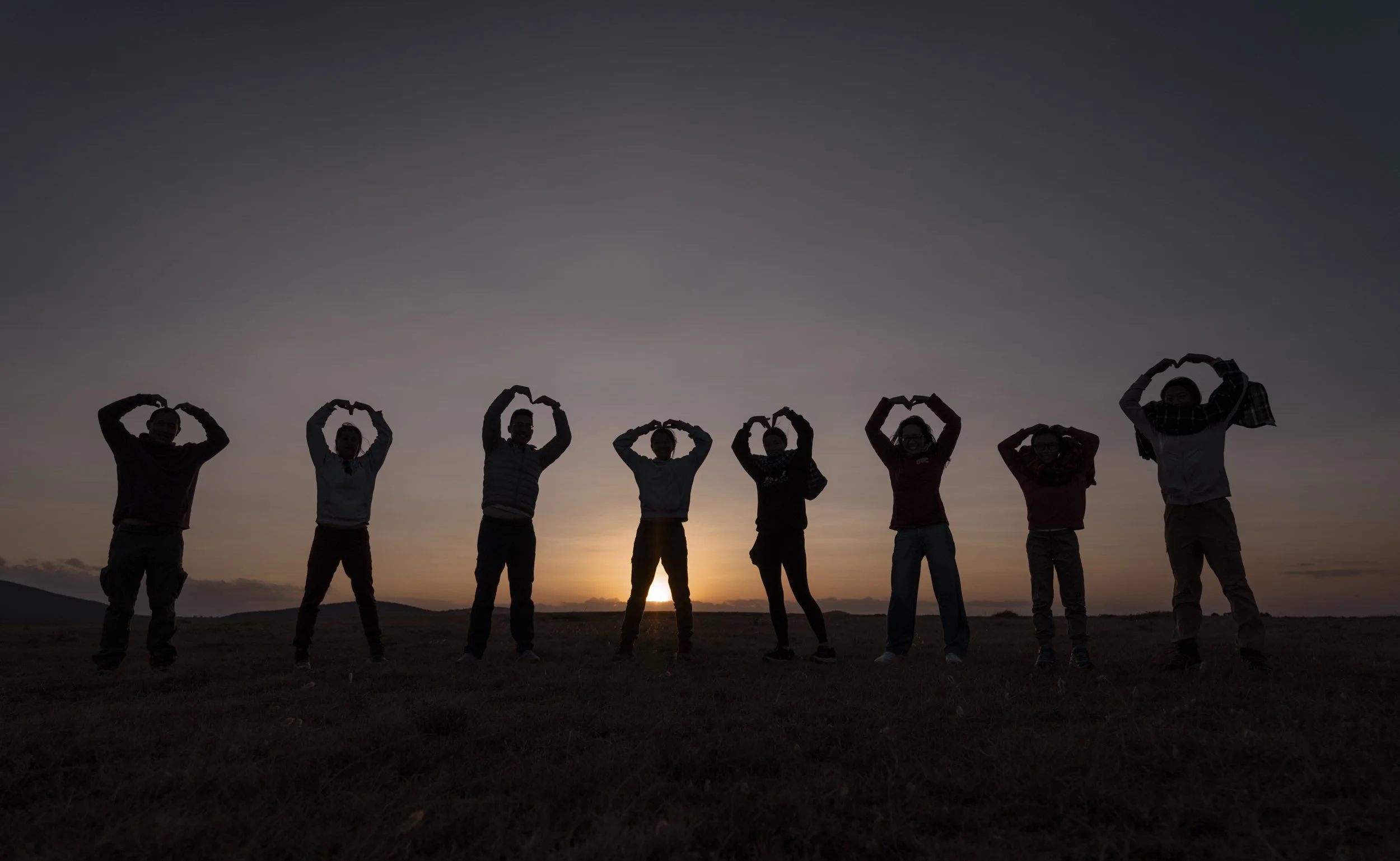 Silhouettes of people forming heart shapes with their arms, standing in a line outdoors during sunset.