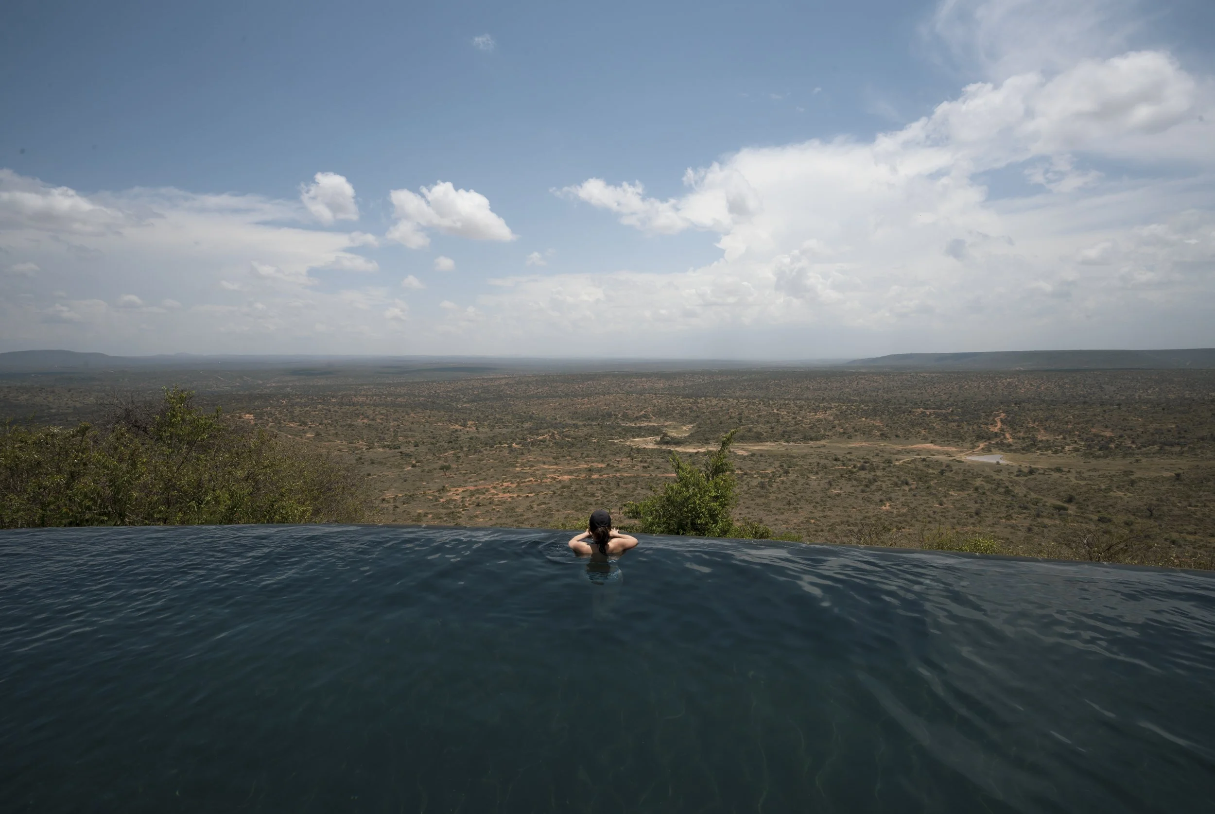 Person relaxing in an infinity pool overlooking a vast arid landscape with sparse vegetation and sky filled with scattered clouds.