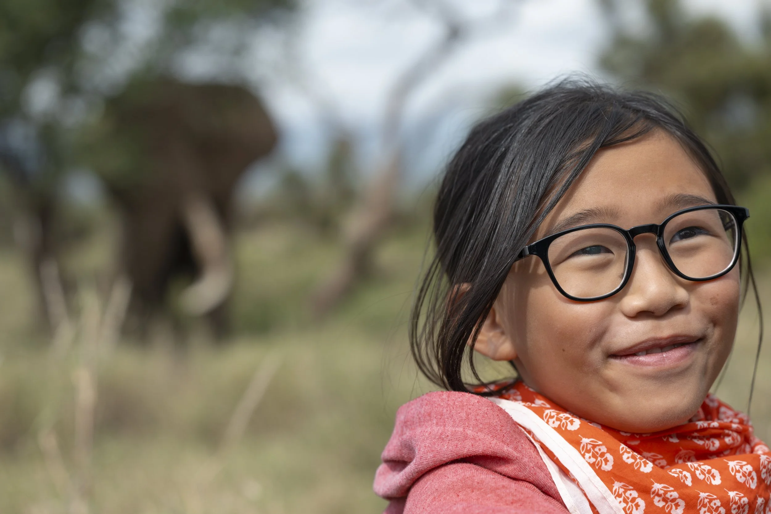 Close-up of a young girl with black hair, glasses, and a bright smile, outdoors with trees in the background.