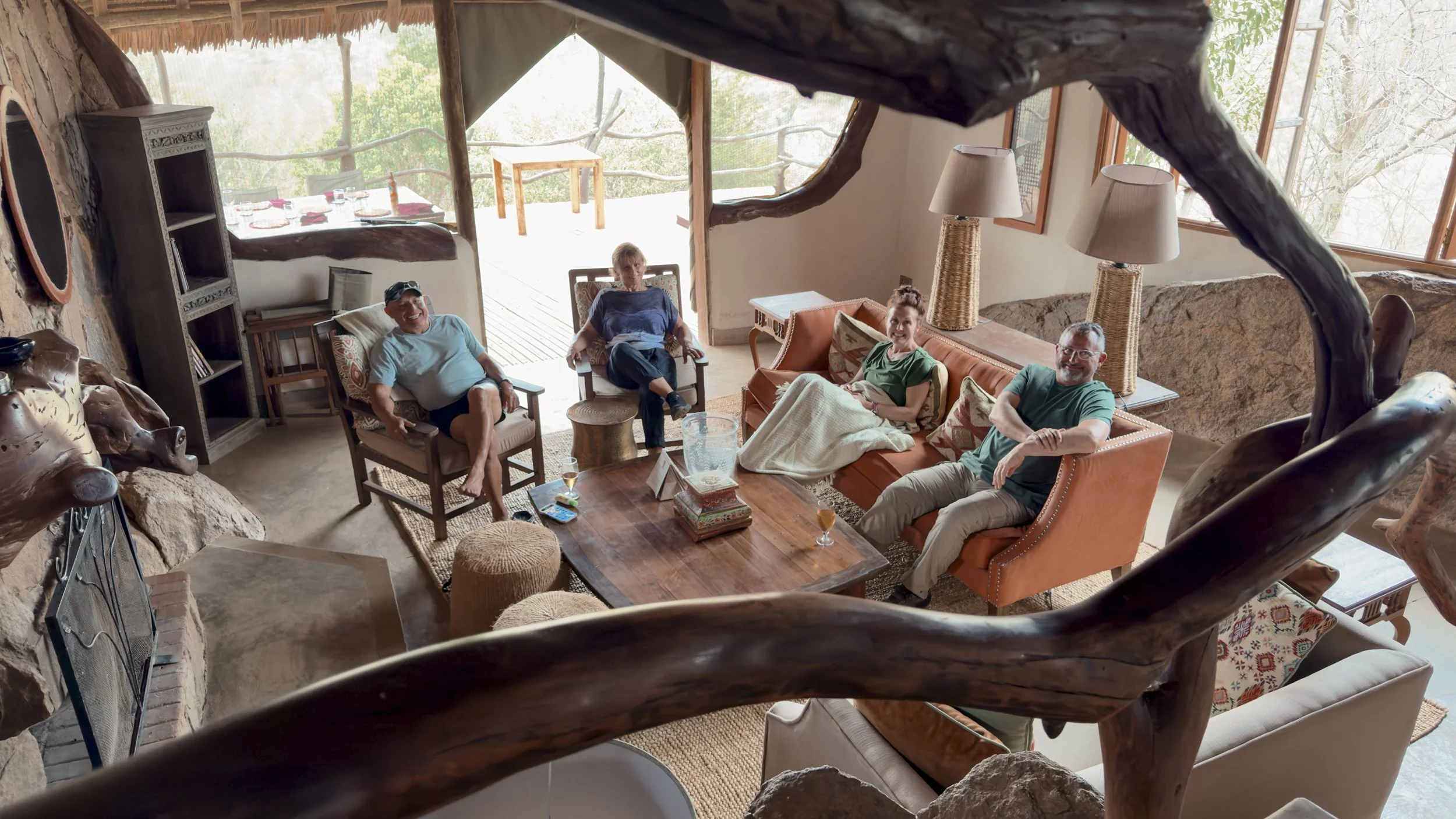 Four people sitting in a living room with large windows, rustic decor, and tree views outside. They are smiling and relaxing, with drinks on the table.