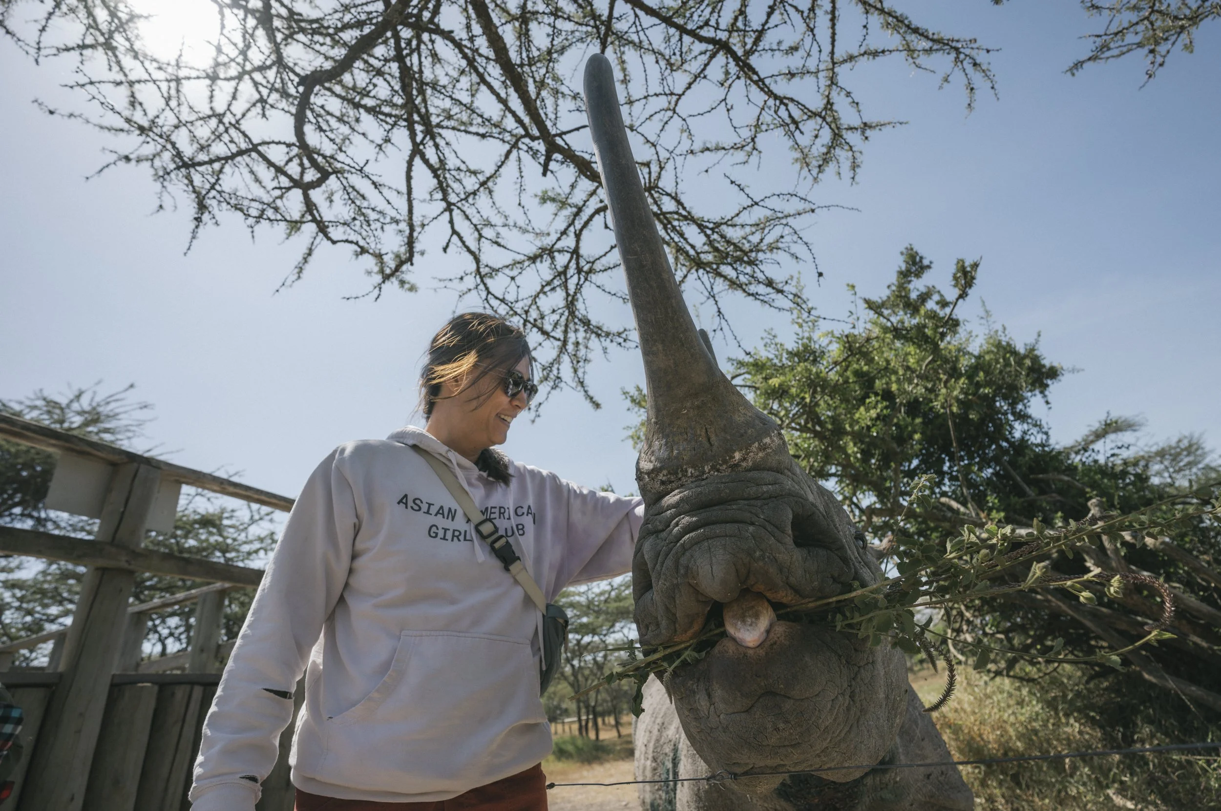 A woman wearing sunglasses and a white hoodie that says 'Asian American Girl Club' is standing outside near a tree with thick branches and a textured trunk. The sky is blue and there are other trees in the background.