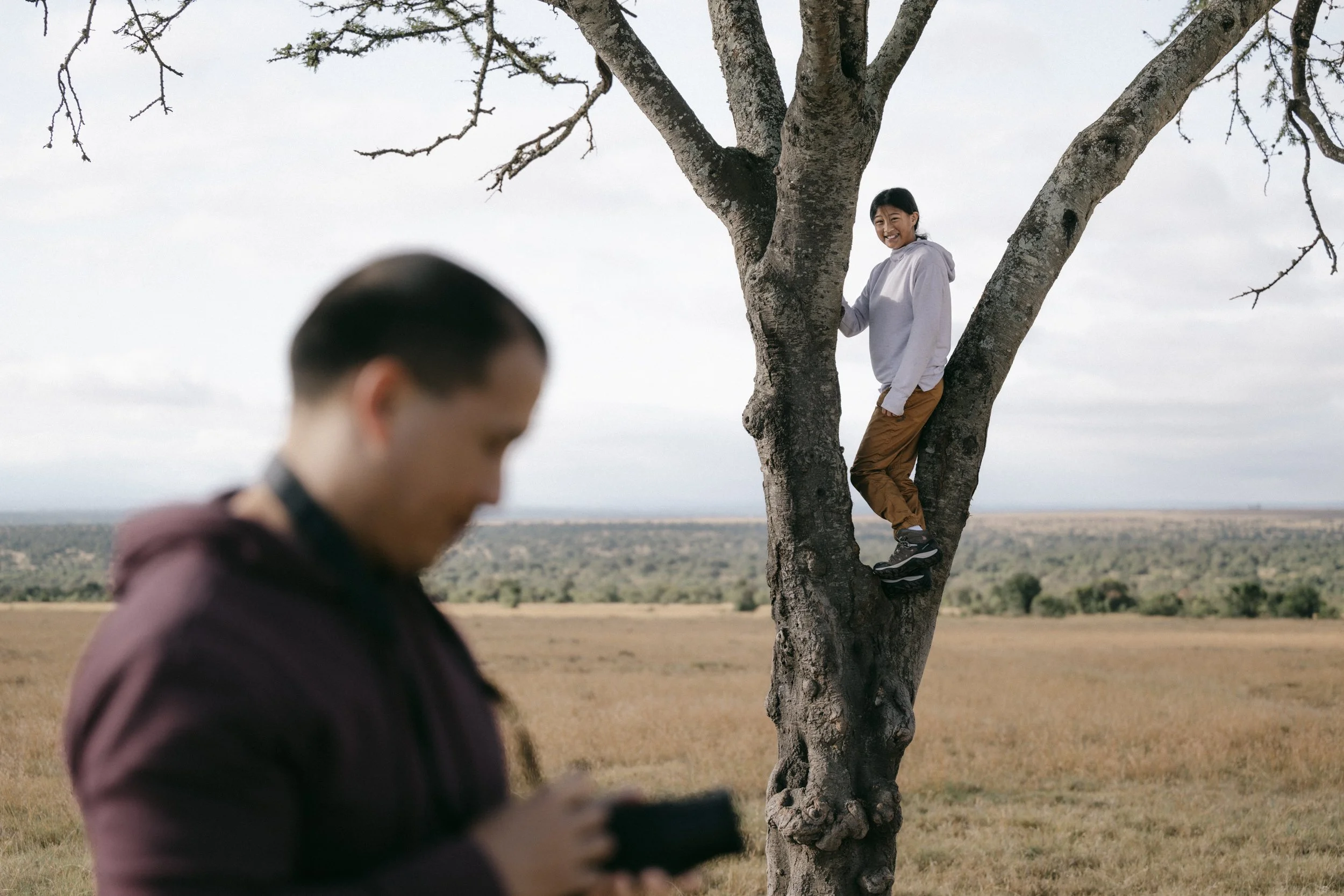 A woman in a light gray hoodie and brown pants standing in a tree with branches, smiling at the camera. In the foreground, a blurred person in a maroon hoodie is looking at their phone. The background shows an open landscape with fields and a cloudy 