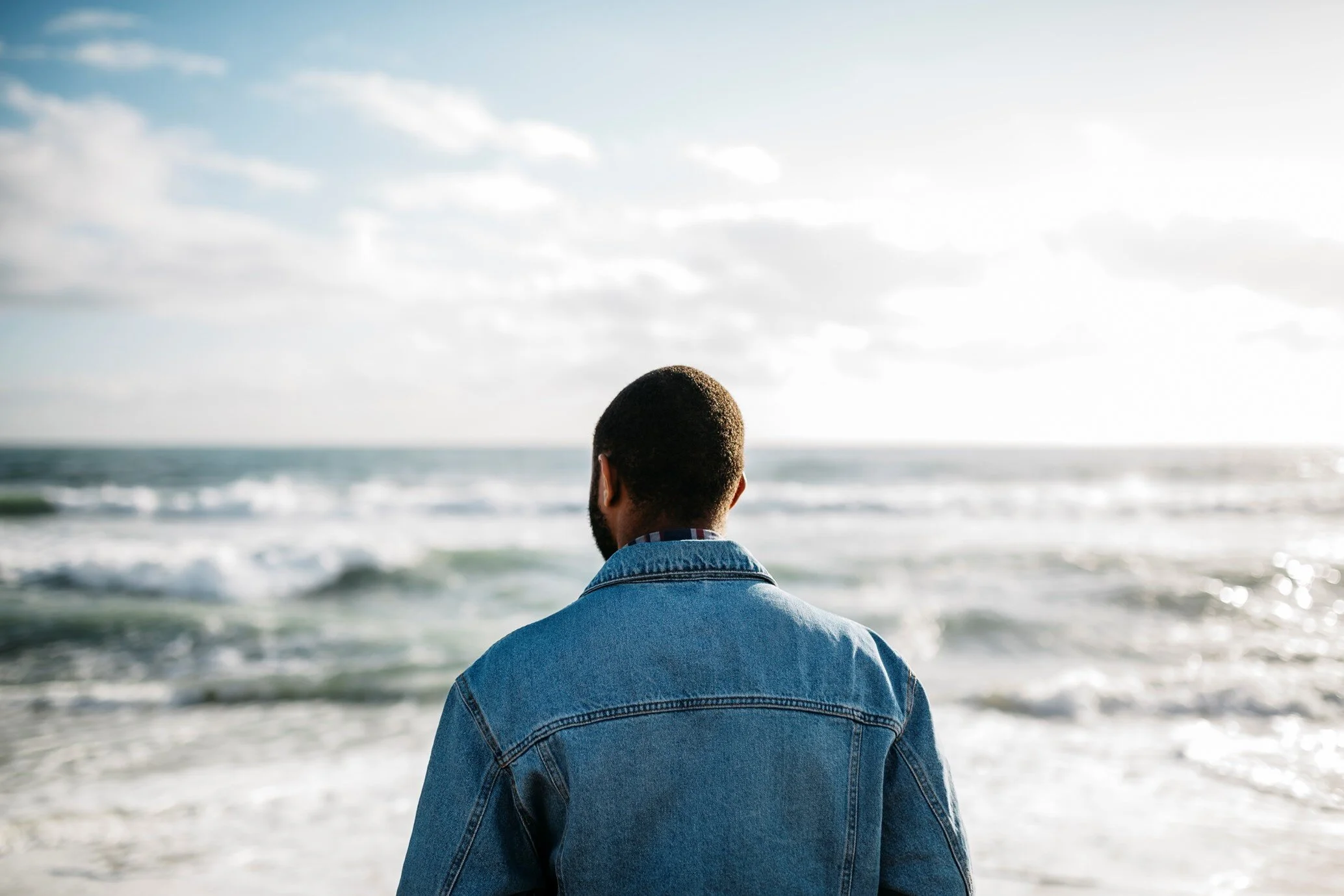 Photo of head and sholders of black man facing the sea.  Back of his head is visible - with his face turned towards the sea. He is wearing a blue denim jacket. In the sky, there are clouds.