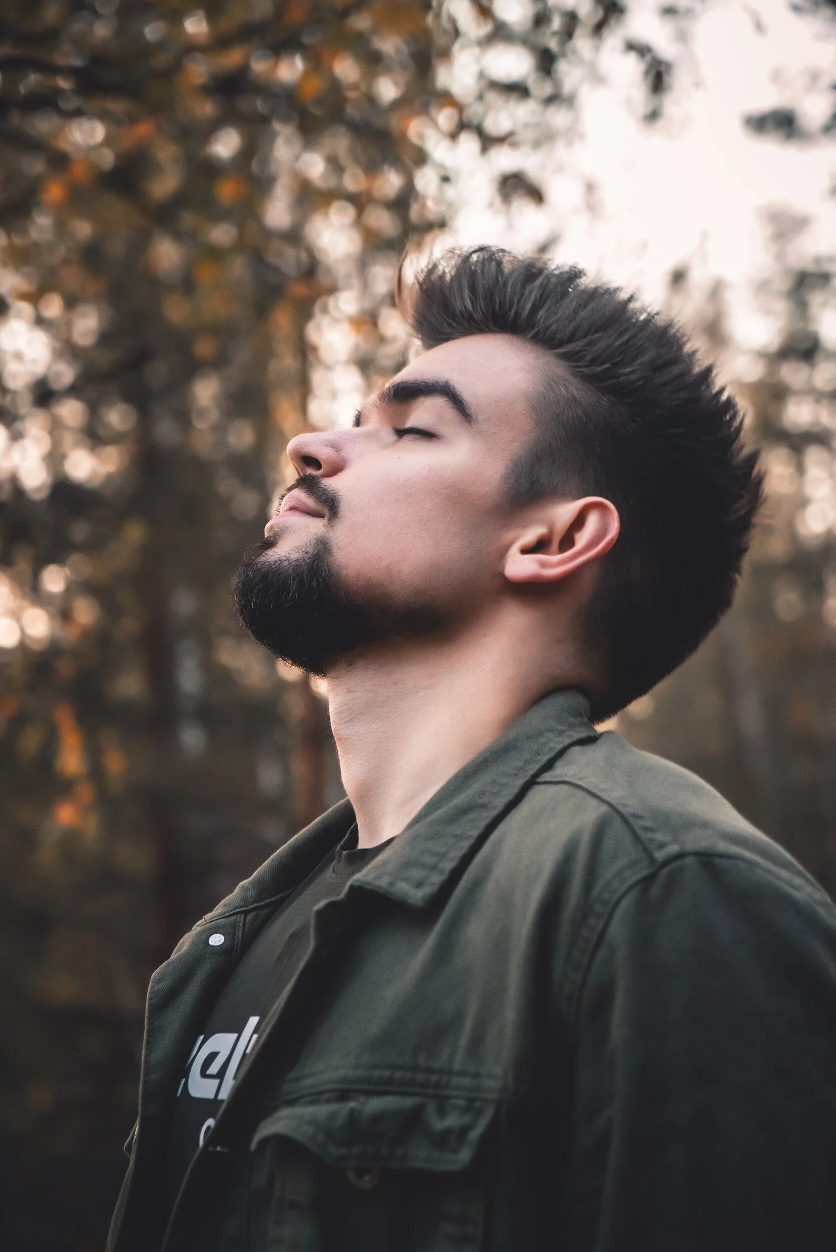 Side profile shoulder and head shot of white man in his 20s stood outside.  He has his eyes closed and head titled upwards.  In the background are trees, somewhat out of focus.