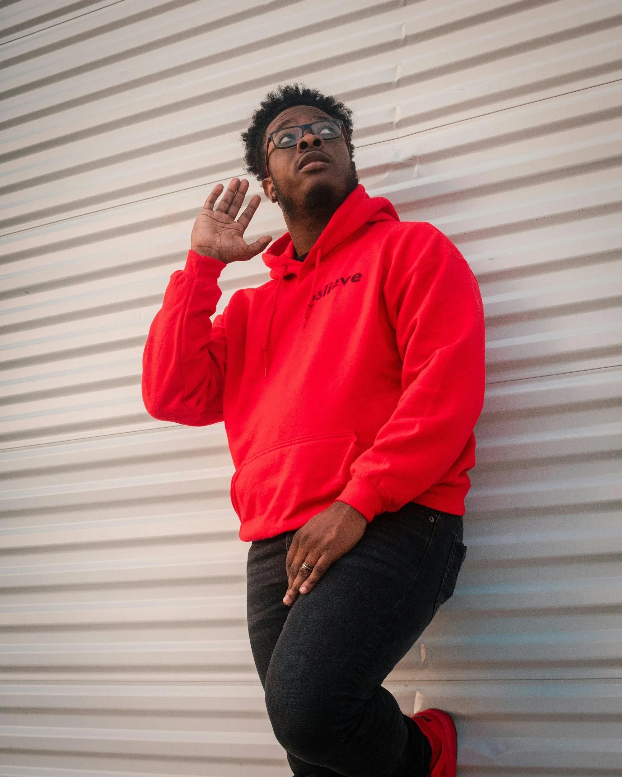 Photo of black man wearing red hoody and looking up. He has his right hand raised up to his ear.