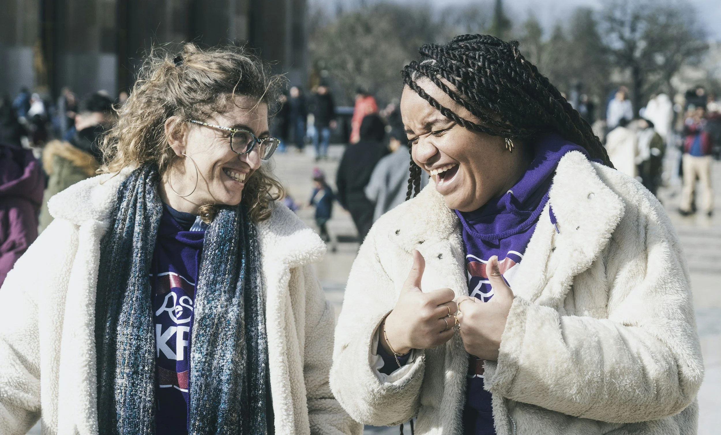 photo of 2 women in a public outdoor space.  They are stood side by side, and are sharing laughter between them - the woman on the left looks at the woman on the right.  The woman on the right has her eyes closed and is laughing