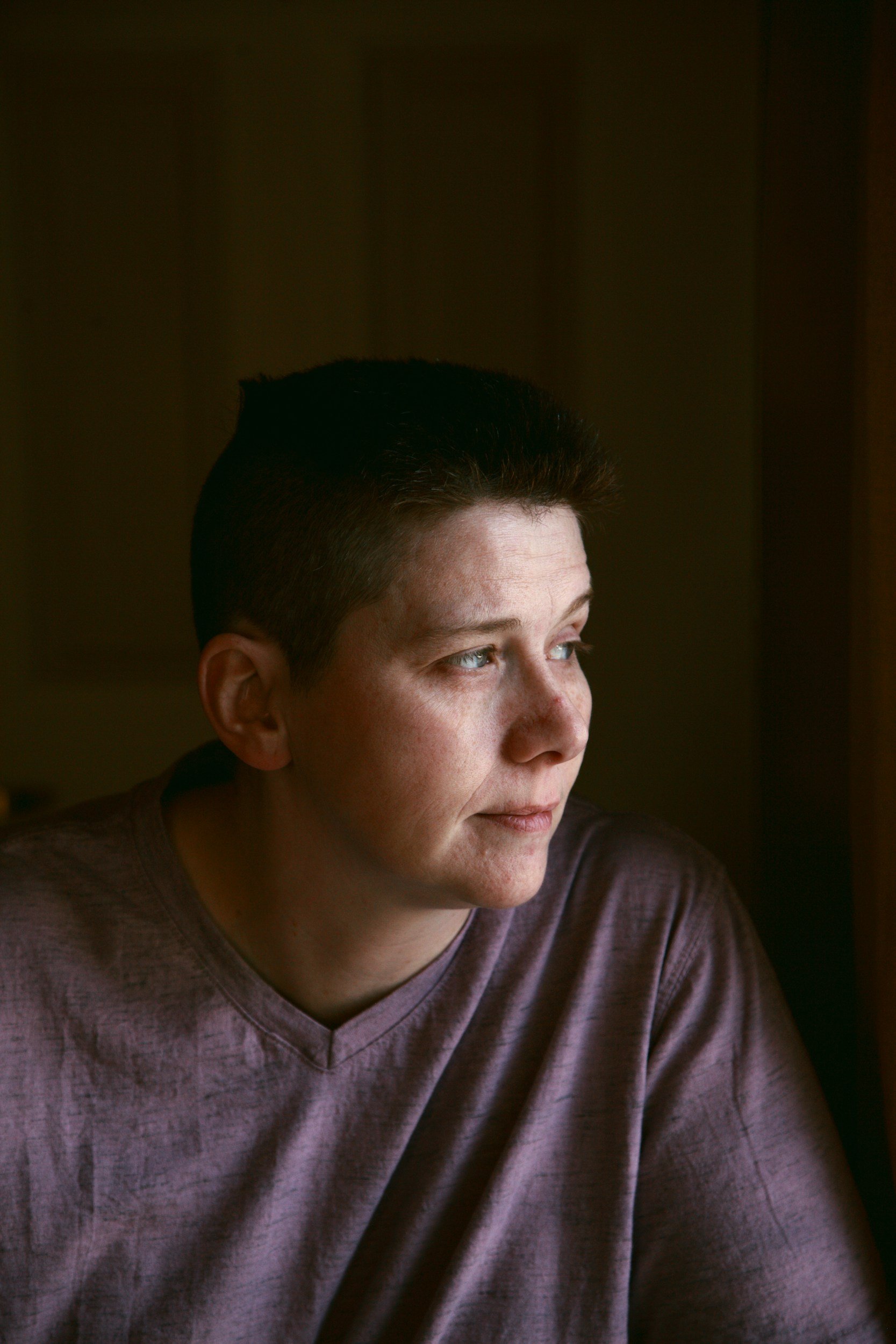 headshot photo taken in lower light conditions. Photo shows a whote person with short dark hair and wearing a grey v-necked t-shirt. They look off to their left with a pensive expression