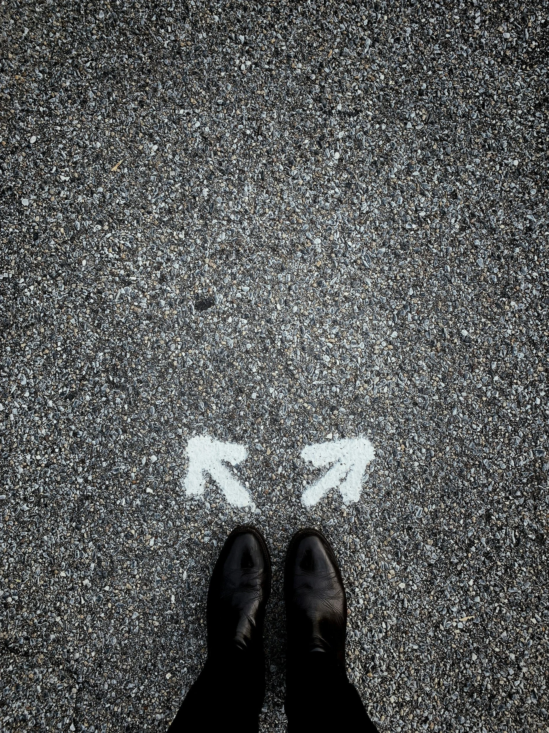 Photo taken from overhead of a person's feet andlower legs - they wear black trousers and black shoes. In front of their feet are two spray painted white arrows (painted onto the gravel ground) pointing in different directions.