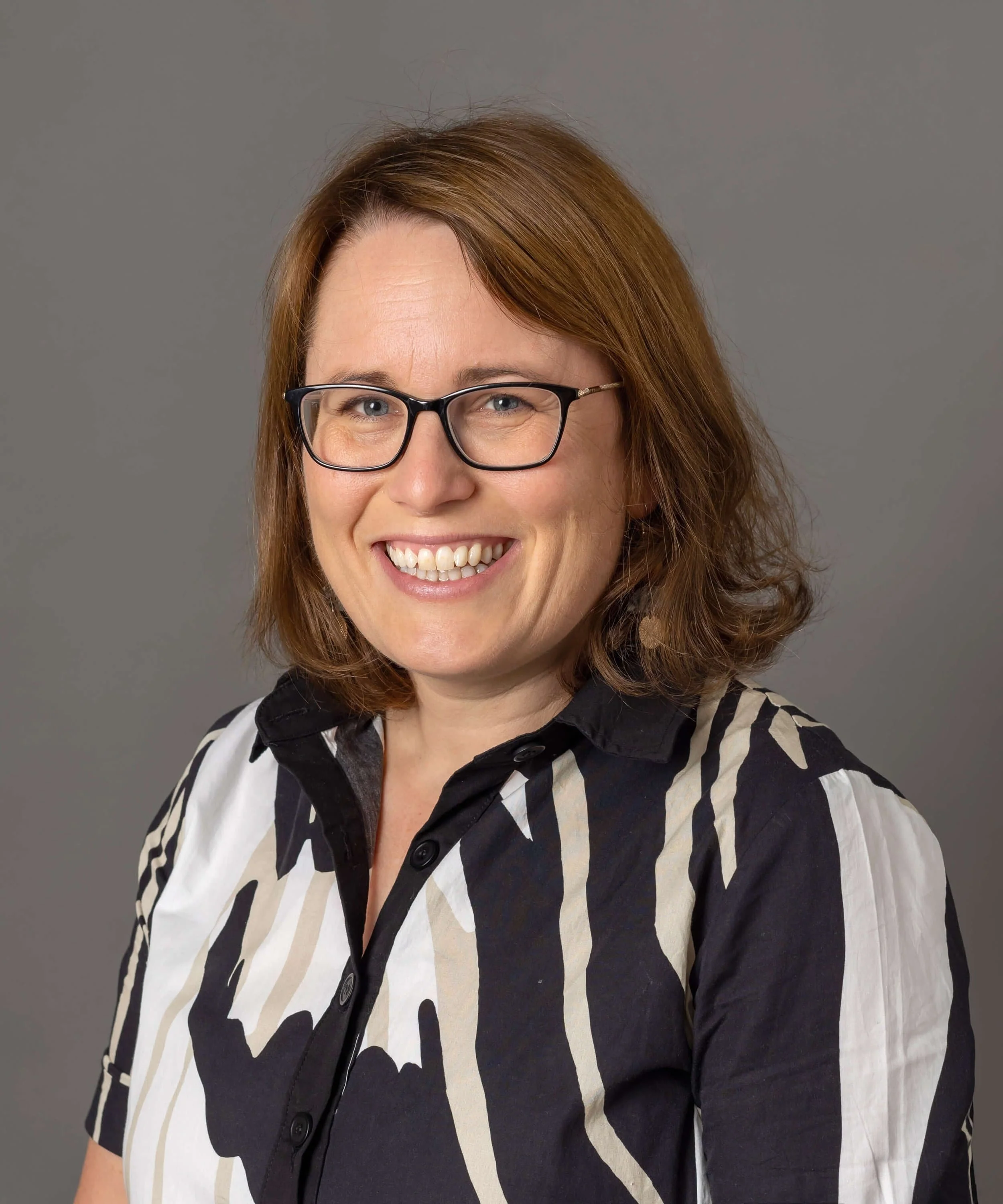 Headshot photo of Claire Law:  she is a white woman with shoulder length brown hair, wearing glasses. She is turned towards the camera smiling. She wears a multi coloured geometric top.