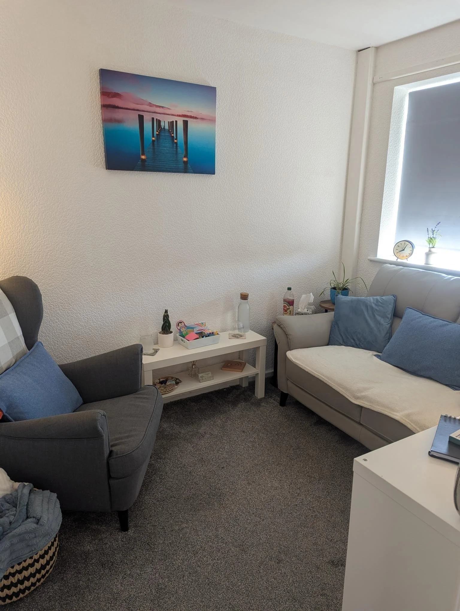 Photo of Claire Law's counselling space - photo has grey wingbacked chair to left of image. opposite grey sofa with blue cushions. There are tissues, water carafe and glass and pot plants and calming picture on the wall