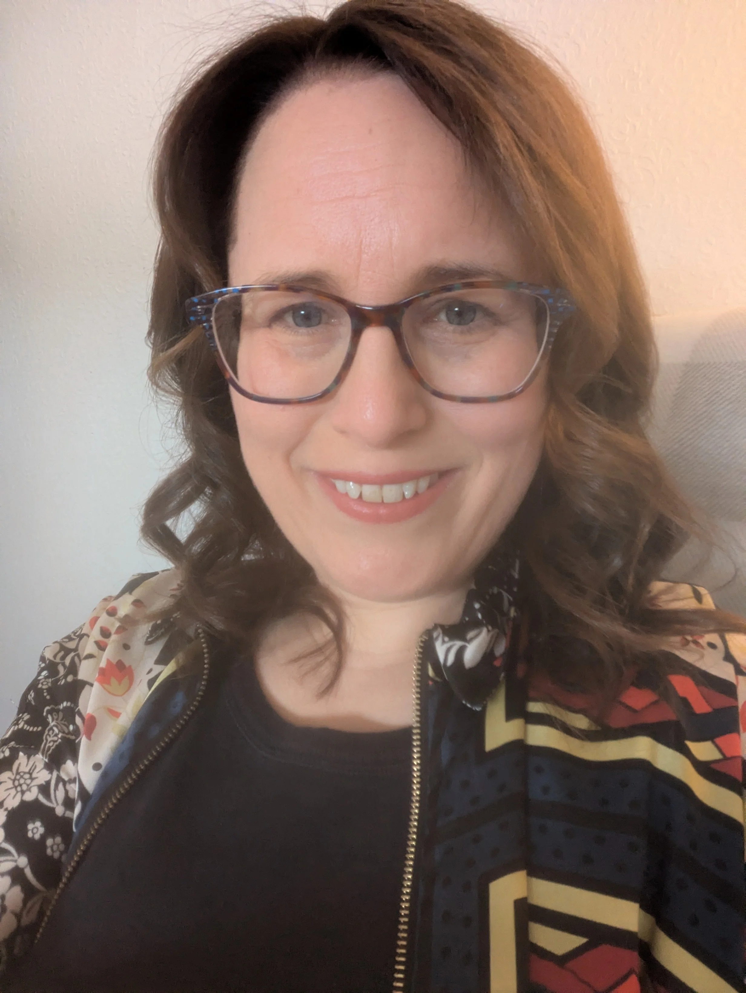Headshot photo of Claire Law:  she is a white woman with shoulder length brown hair, wearing glasses. She is turned towards the camera smiling. Claire wears a colourful geometric open short over a navy top