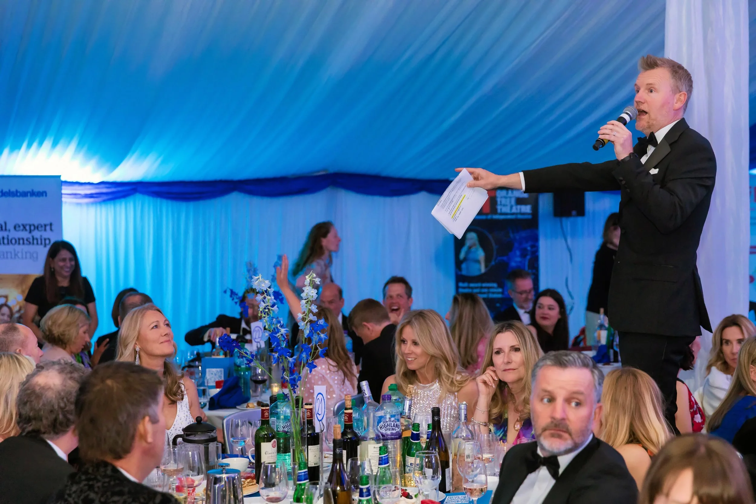 A compere in black tie standing on a chair announcing prize winners at a charity ball