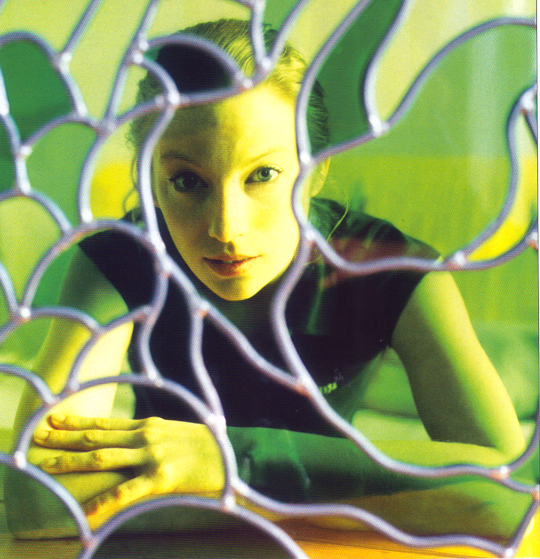 A woman looking through a decorative wire and green-painted holes at the camera.