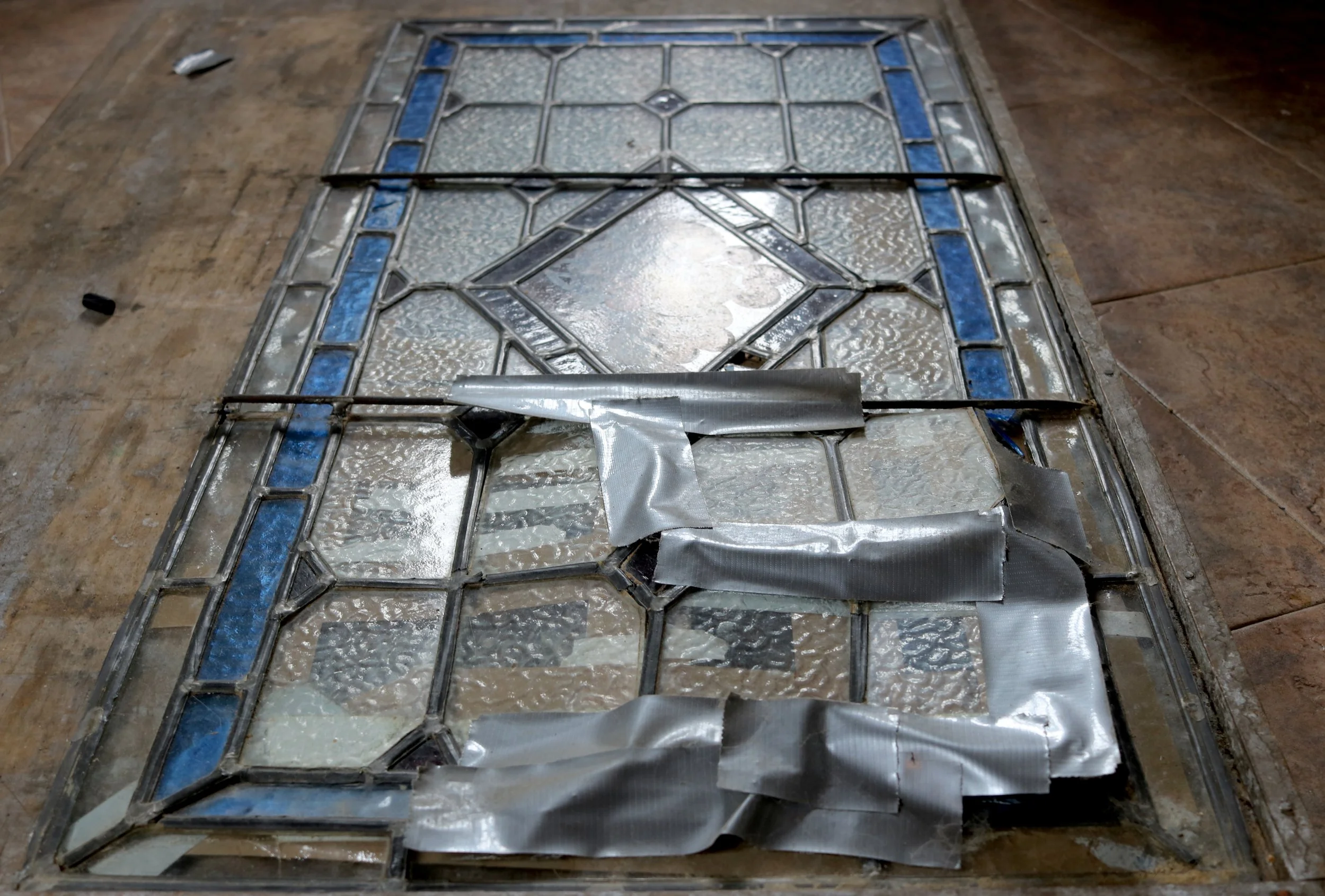 Two stained glass window panels lying on a floor with tools around them, one panel stacked on top of the other and partially covered in protective plastic film