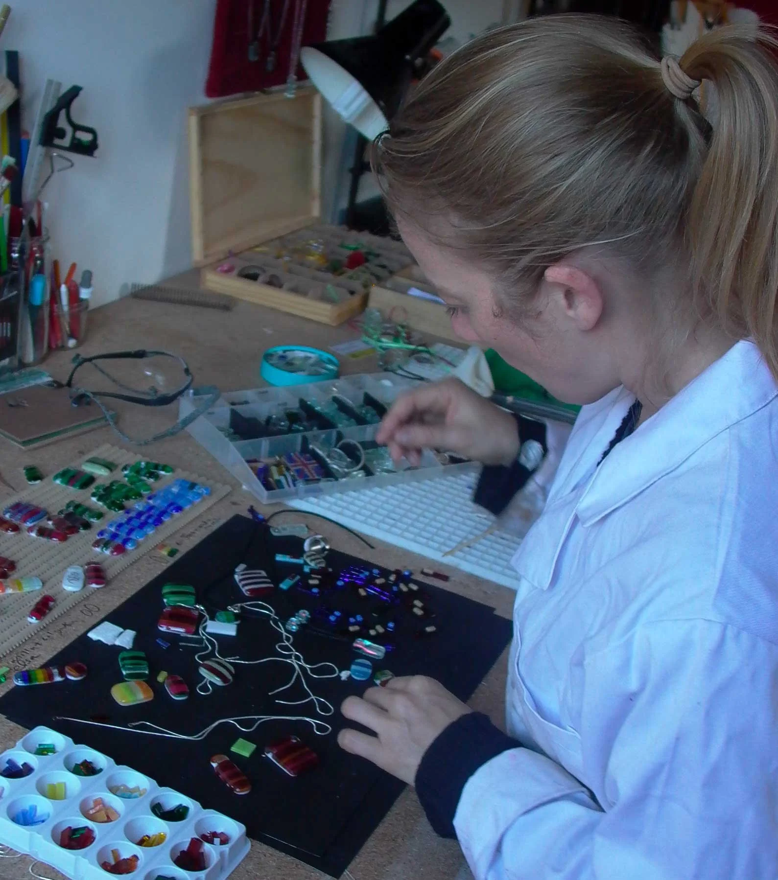 A woman working on jewelry or small craft projects at a cluttered workbench, with various colorful pieces and tools spread out across the surface and organized in small containers.