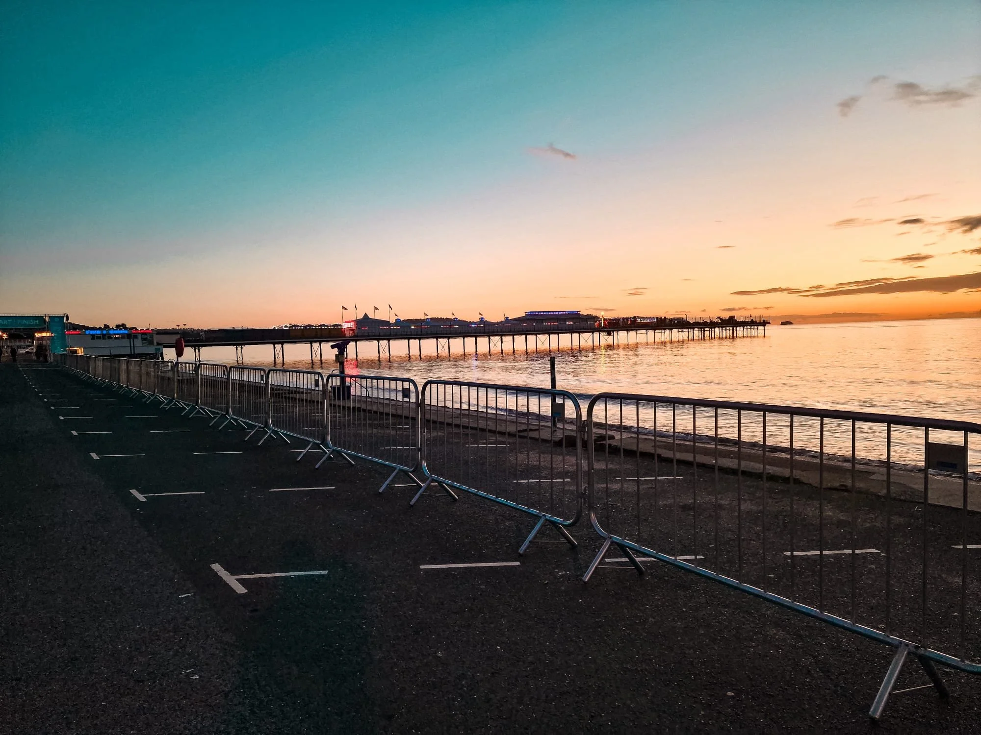 A pier extending over calm water during sunset with colorful sky, some clouds, small buildings and flags along the pier, and empty parking lot with barricades in foreground.