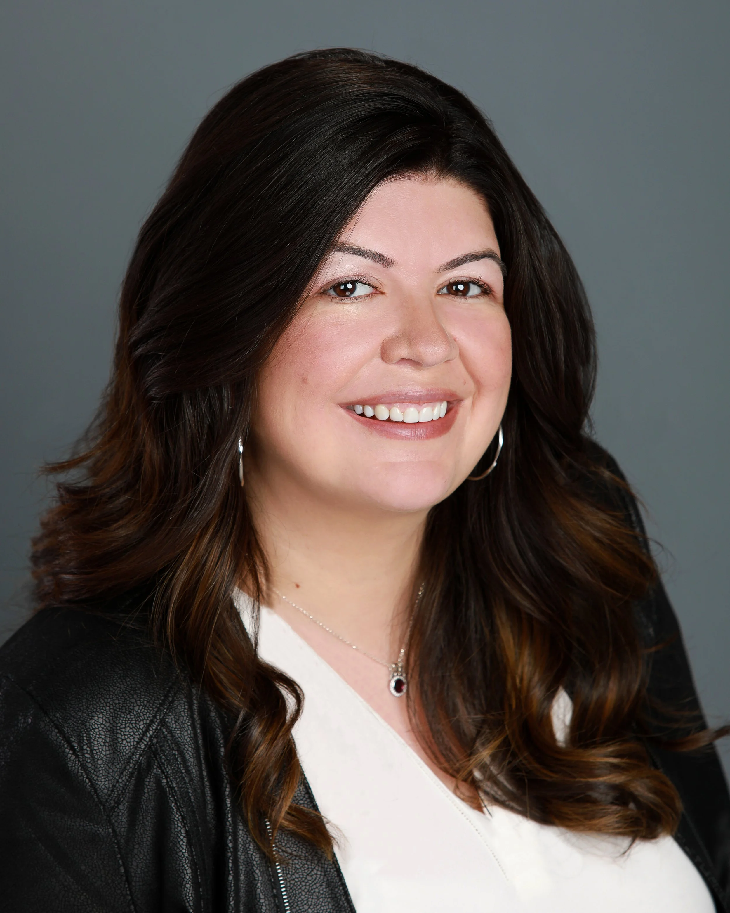 Close-up of a smiling woman with long dark wavy hair, wearing a white top, black jacket with leather texture on the sleeves, hoop earrings, and a necklace with a red gemstone pendant, against a gray background.
