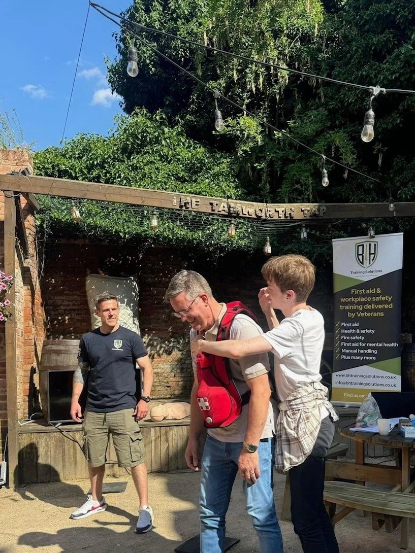 Three men in an outdoor setting with string lights overhead. One man is adjusting a first aid kit on another man's chest. A third man watches nearby. There is a sign behind them about workplace safety training, and a table with various items, including a laptop and cups.