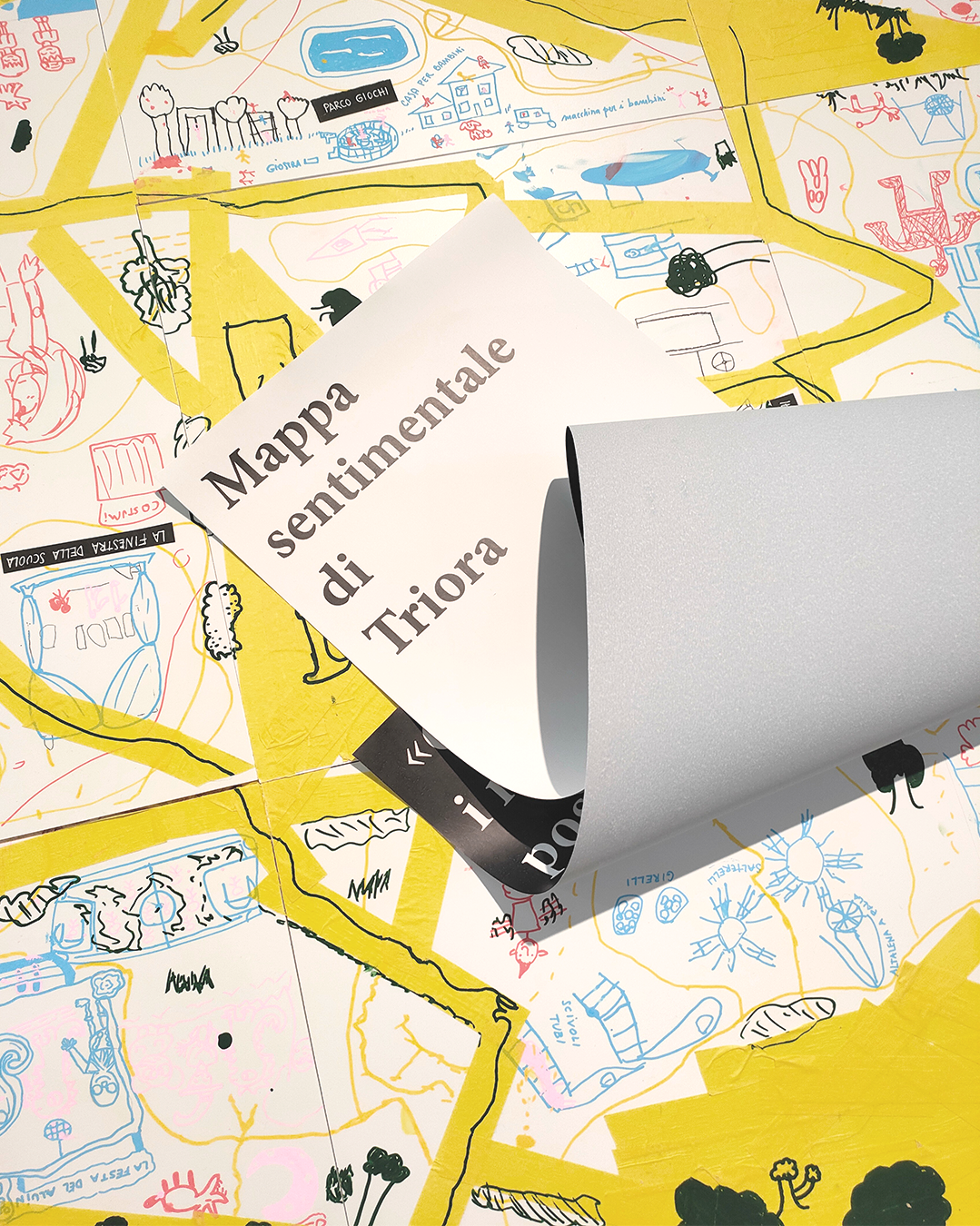 Mappa sentimentale di Triora su una superficie ricoperta di mappe e disegni colorati, con una pagina di carta bianca che si solleva.