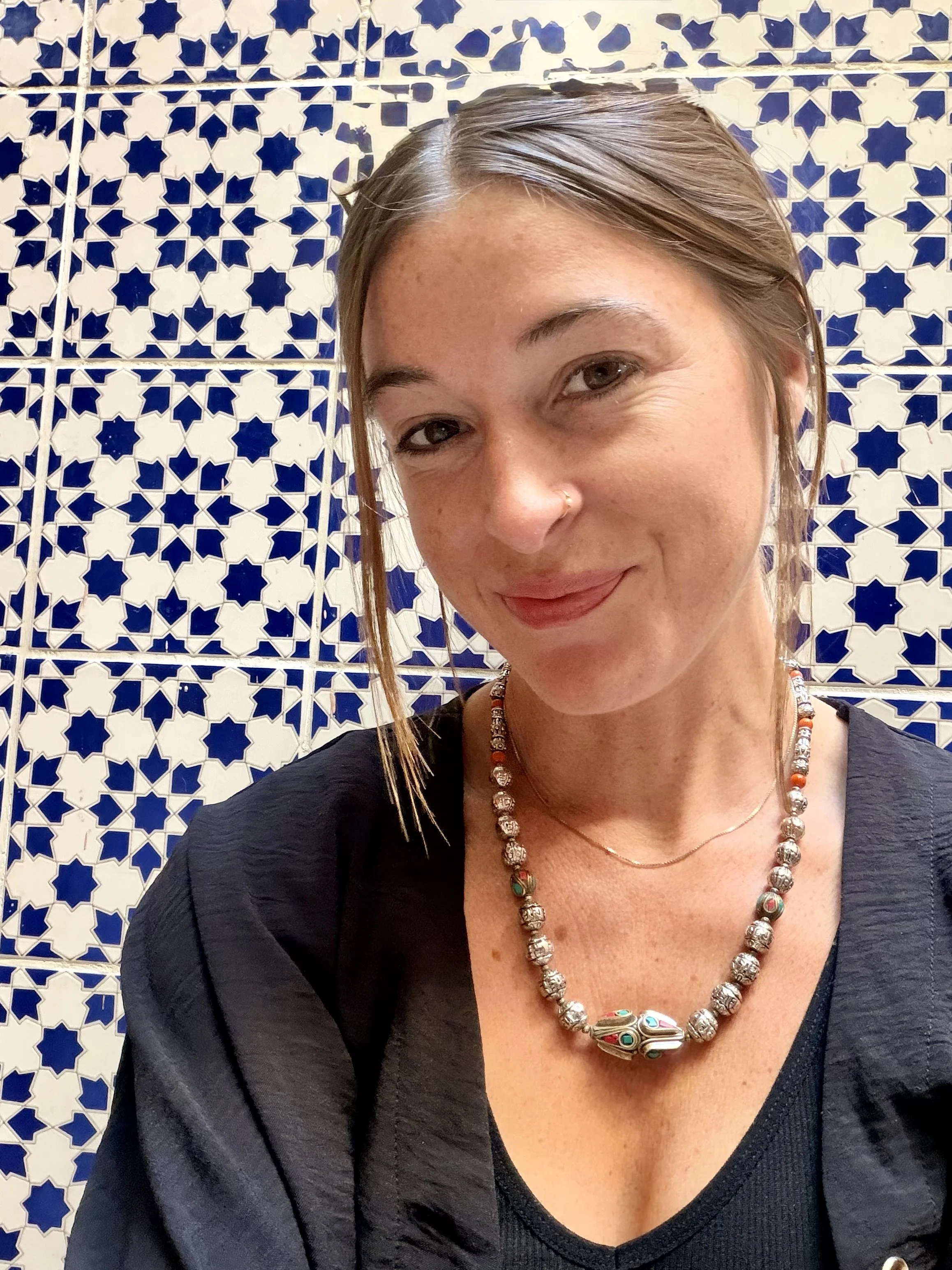 Smiling woman with dark hair and floral dress outdoors in a nature setting with trees and cliffs in the background.