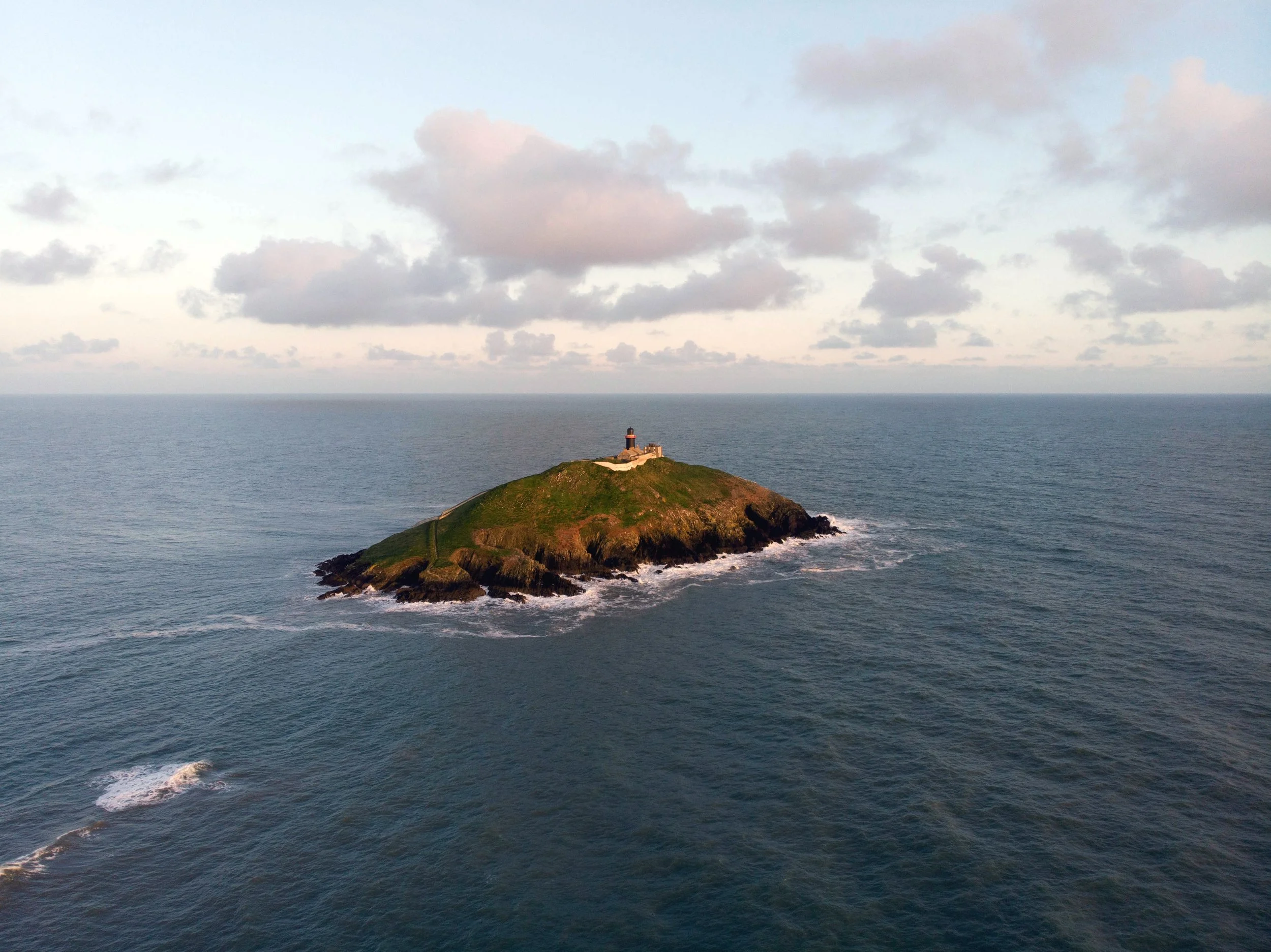 An aerial view of a small island with a lighthouse, surrounded by open sea, part of the natural imagery of The Oya Energy in Colchester and online.