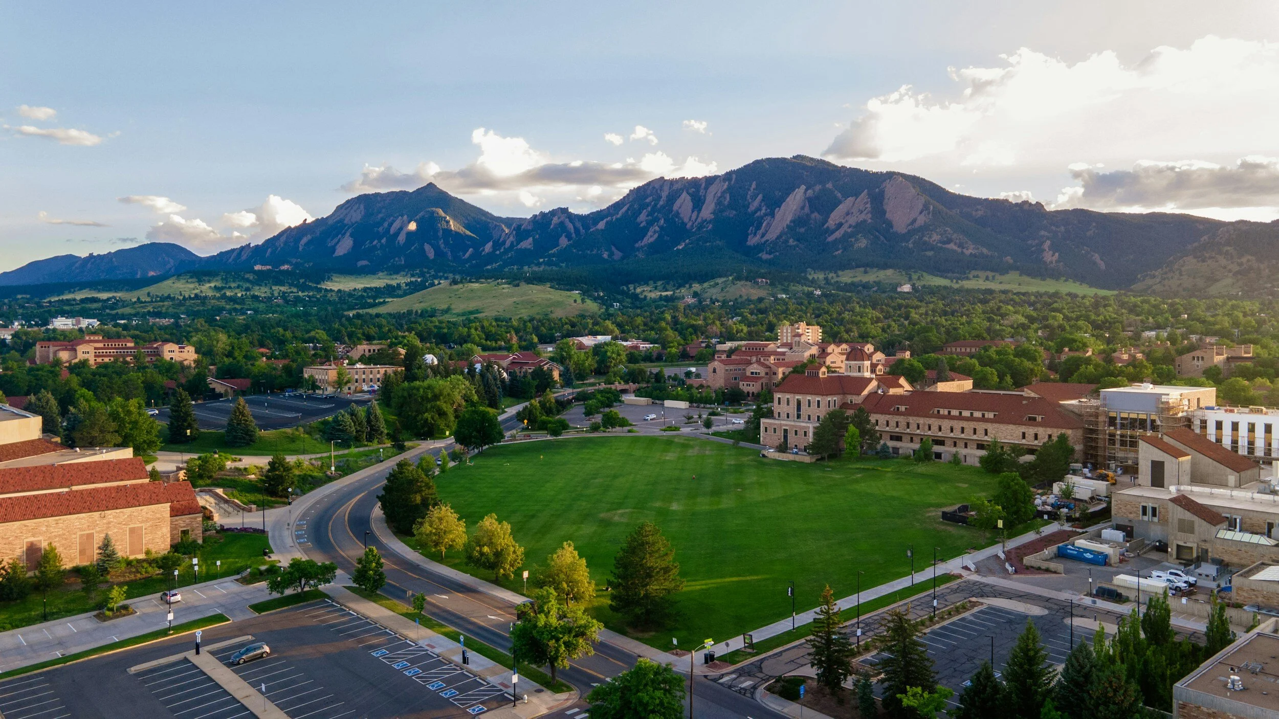 Aerial view of a college campus with grassy fields, trees, and historic buildings, with mountains in the background under a partly cloudy sky.