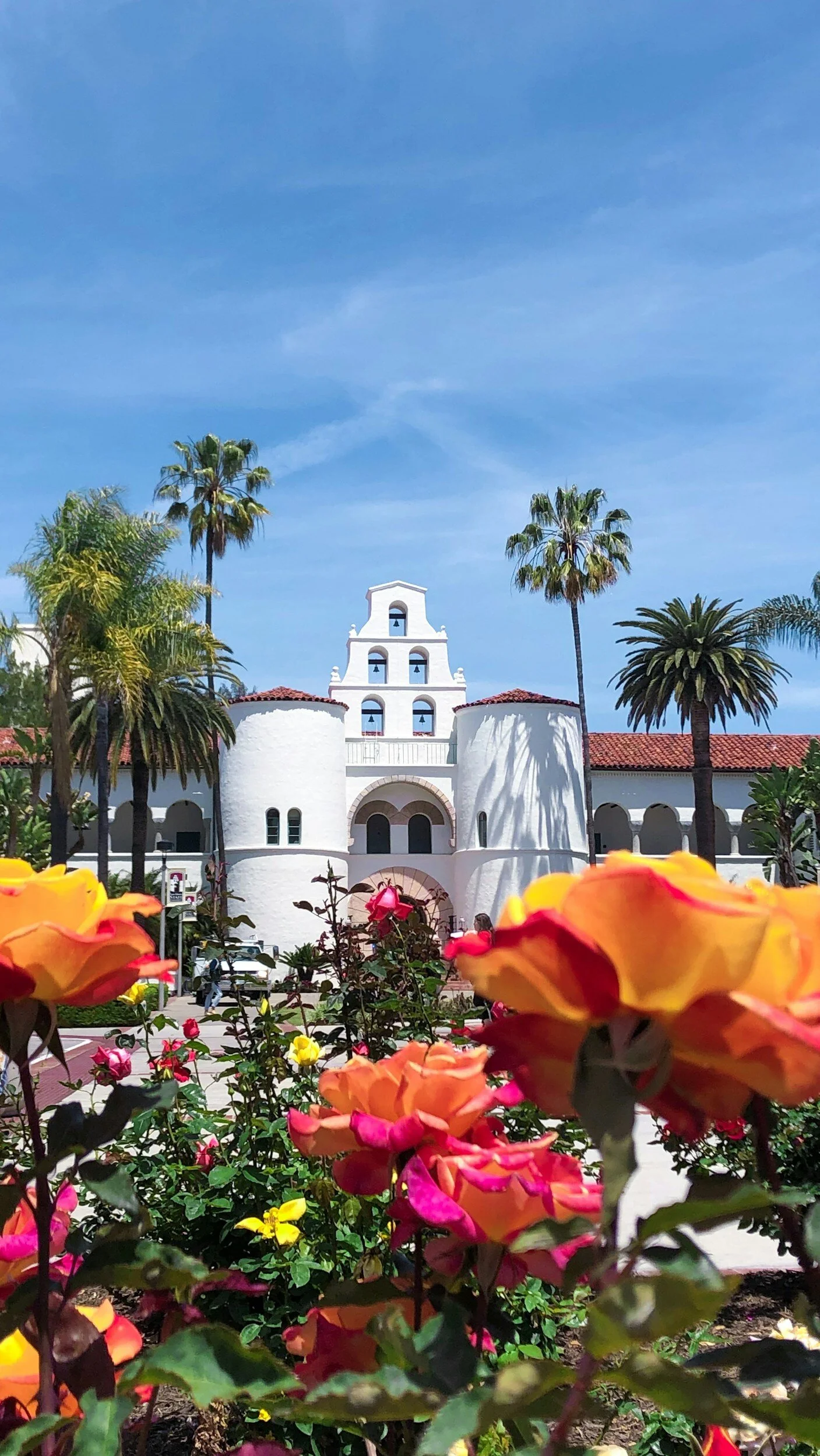A white Spanish-style building with a bell tower, surrounded by palm trees and colorful flowers, under a bright blue sky.