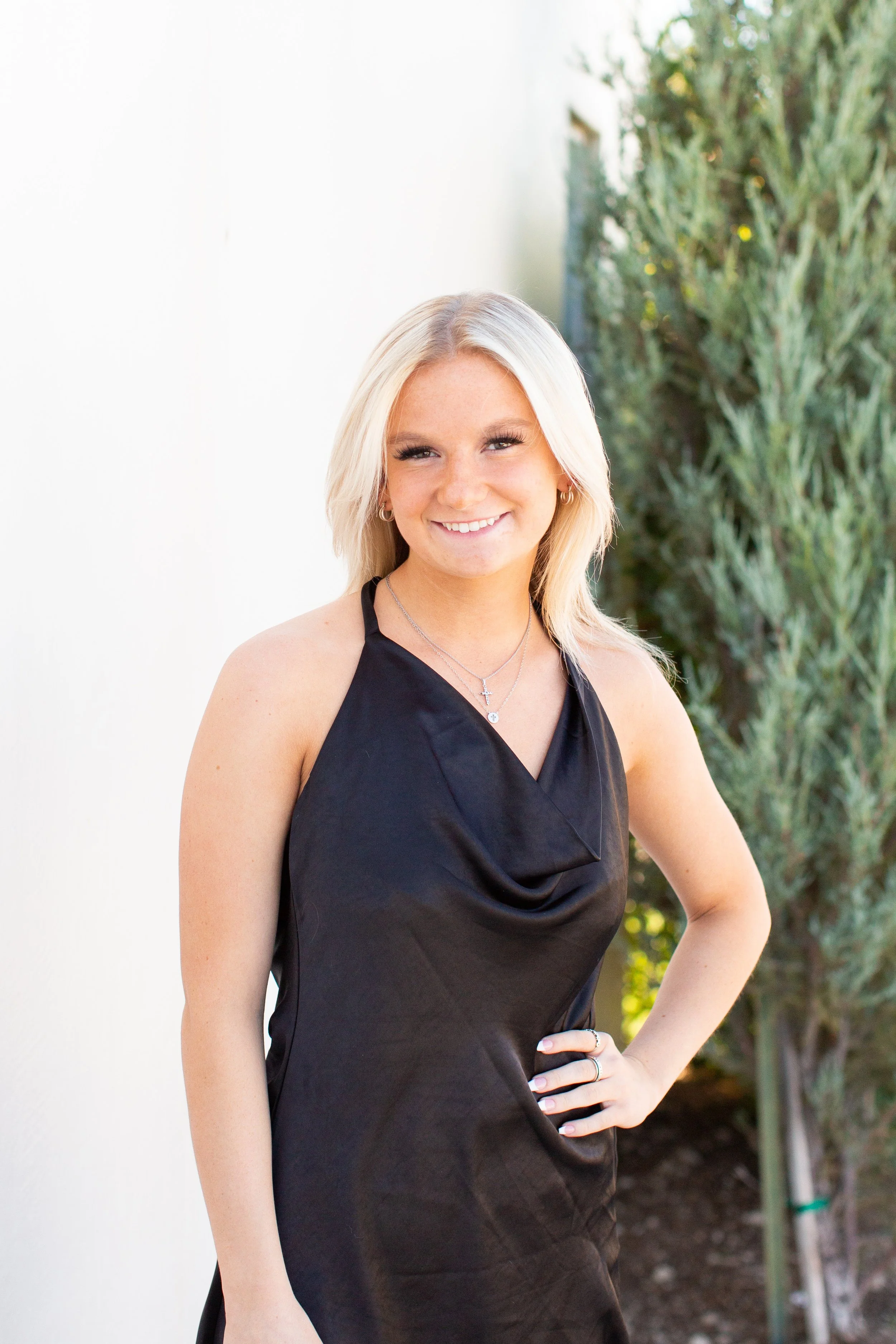 A young woman with blonde hair wearing a black sleeveless dress, standing outdoors, smiling, with greenery in the background.