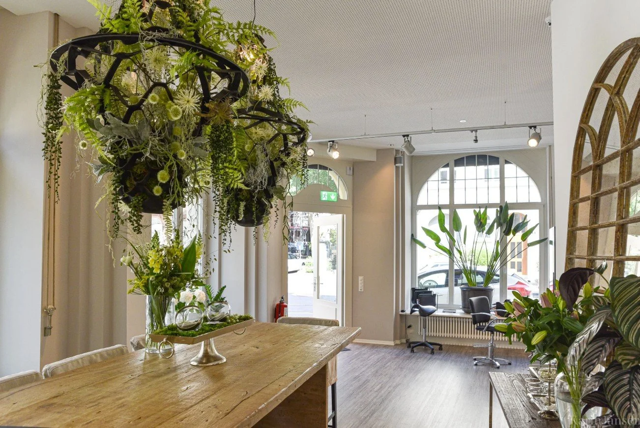 A modern hair salon interior with beige styling chairs on wooden floors, a central white station with mirrors, and natural light coming through two windows.