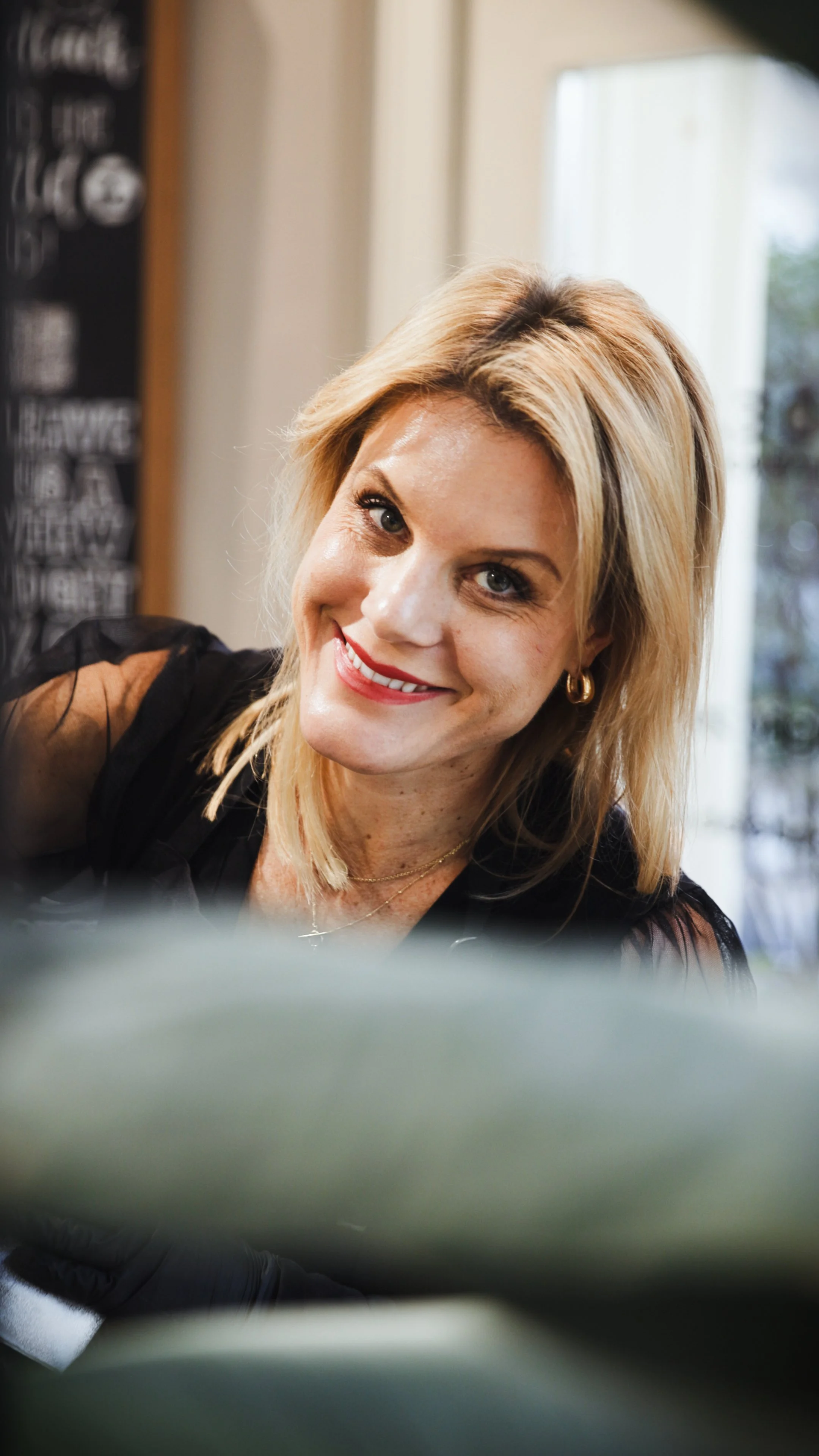 A woman sitting against a gray background, wearing a black sleeveless top and black pants, smiling with blonde hair styled in soft waves.