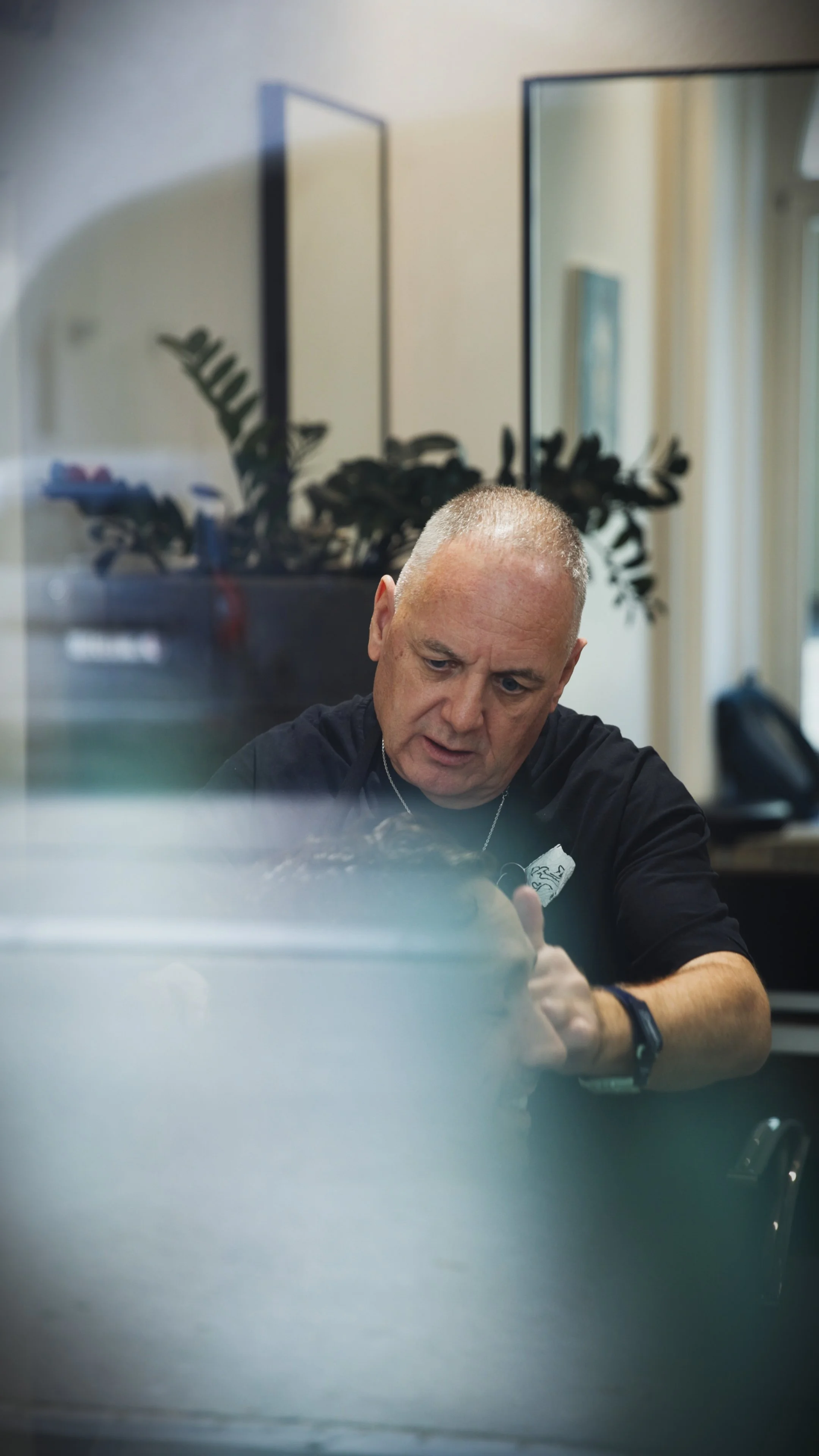 A middle-aged man with short gray hair, wearing a white t-shirt with pocket details, black pants, a wristwatch, and bracelets, sitting on a wooden stool against a gray background.