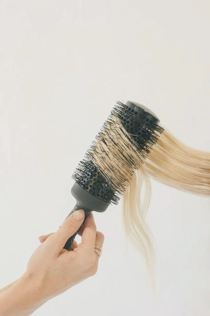 A hand holding a round hairbrush with black bristles and blonde hair wrapped around it against a plain white background.