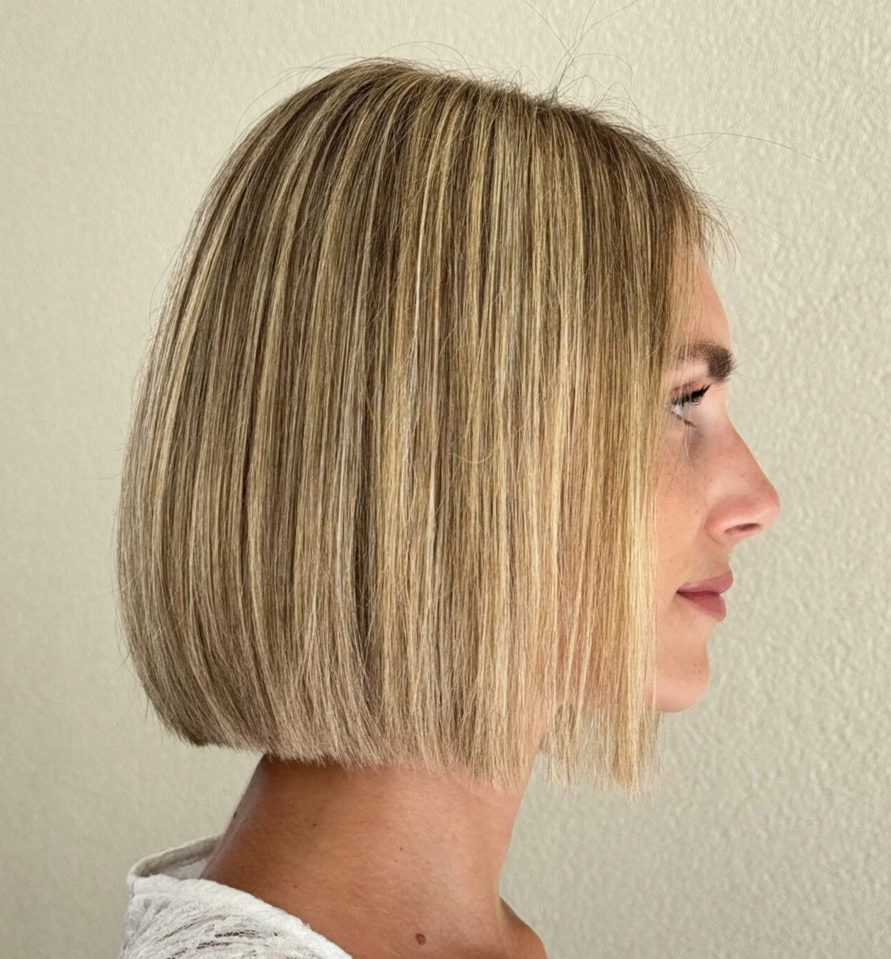 Side view of a woman with shoulder-length blonde hair and subtle makeup, standing against a plain wall.