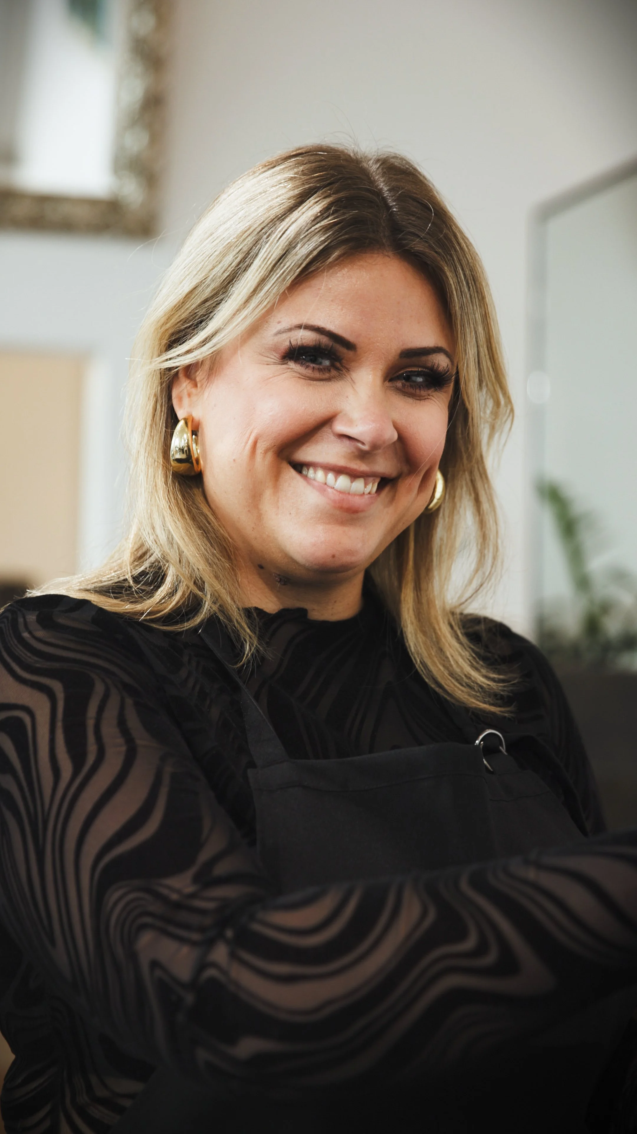 A woman with long blonde hair, wearing a black satin jacket and lace top, sitting against a gray background, smiling at the camera.