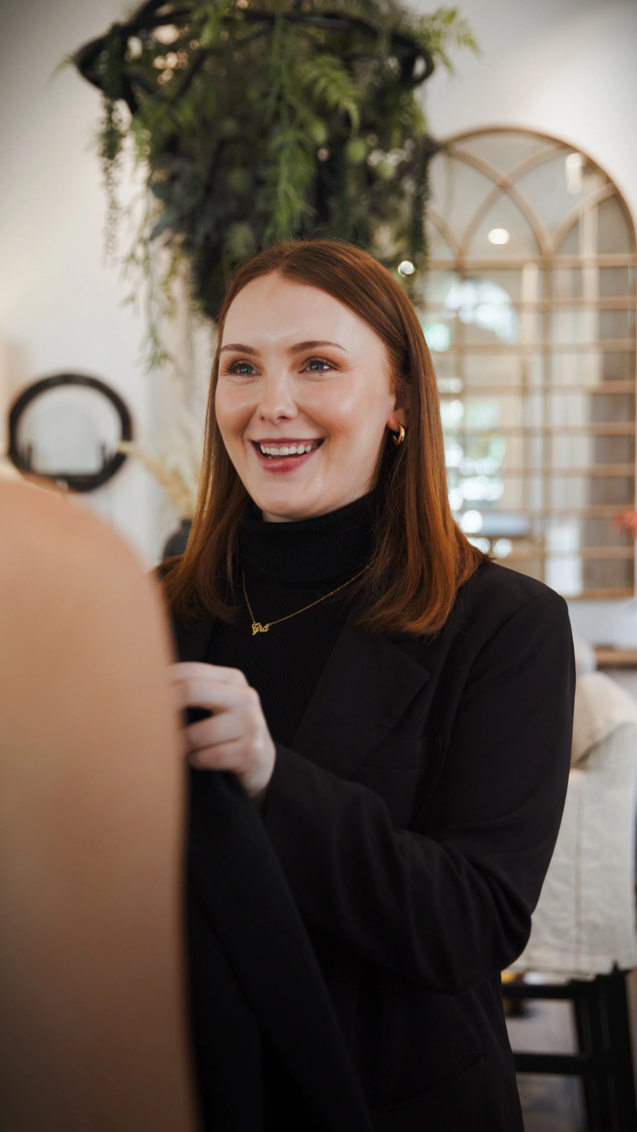 A young woman with long red hair and a bright smile, wearing a black top with ruffled sleeves and black pants, sitting on a wooden box against a plain gray background.